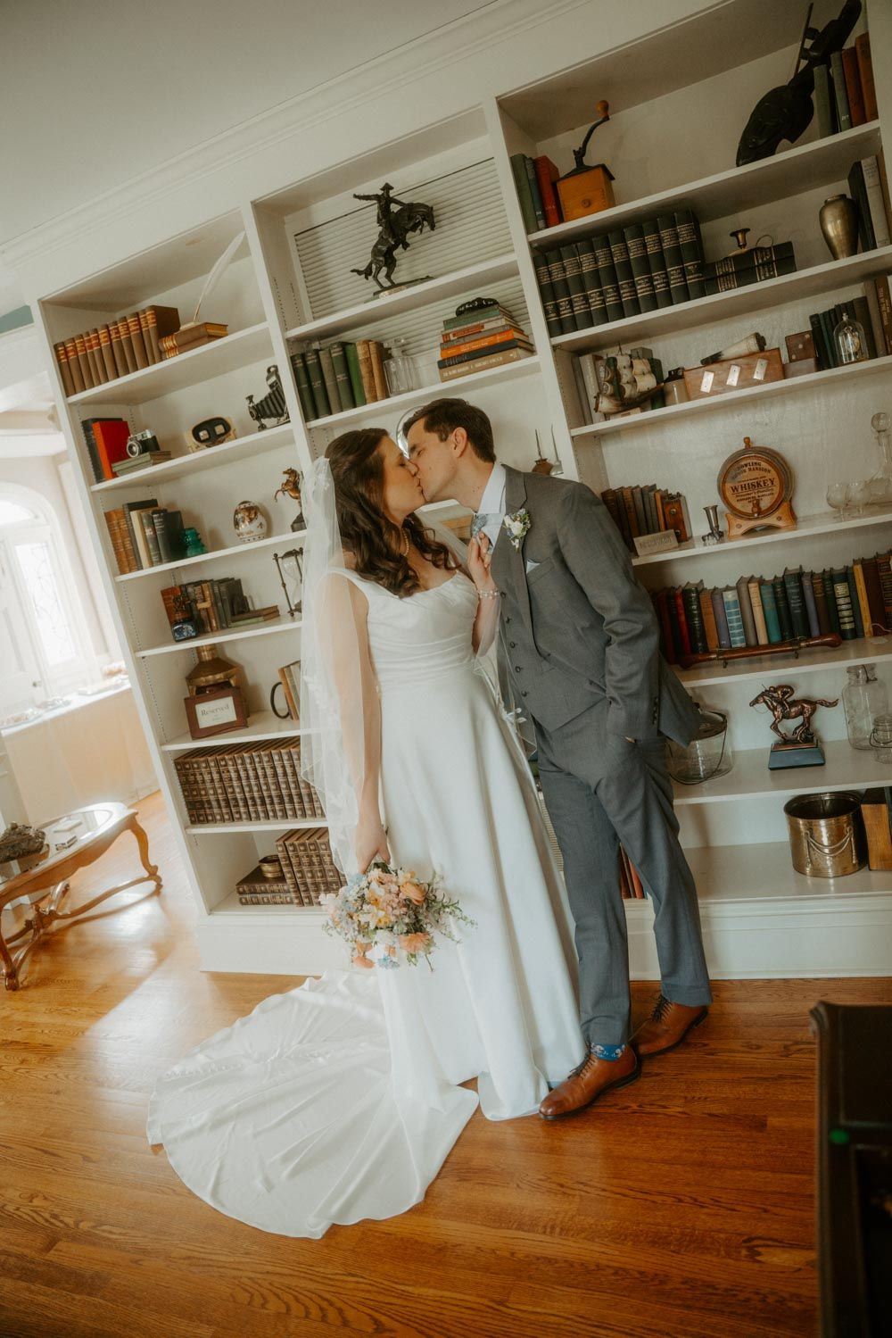 Bride and groom share a kiss in a charming library room with wooden floors and book-filled shelves on their wedding day.
