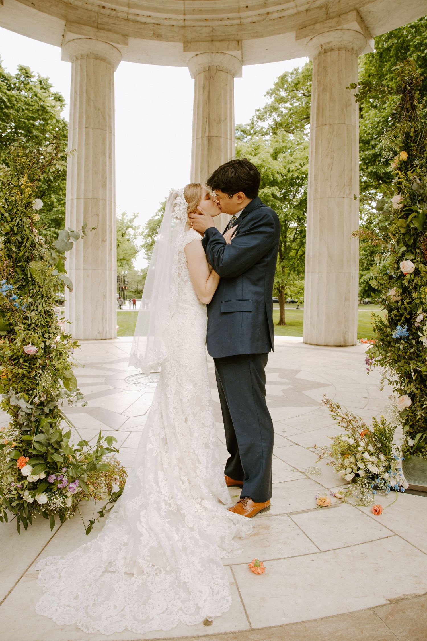 Bride and groom share first kiss under a floral-adorned marble pavilion during an elegant outdoor wedding ceremony.