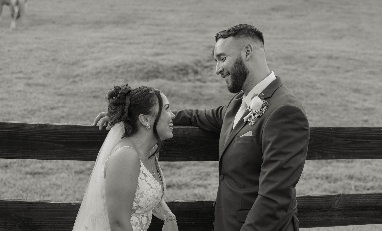Black and white photo of bride and groom sharing a joyful moment by a rustic wooden fence.