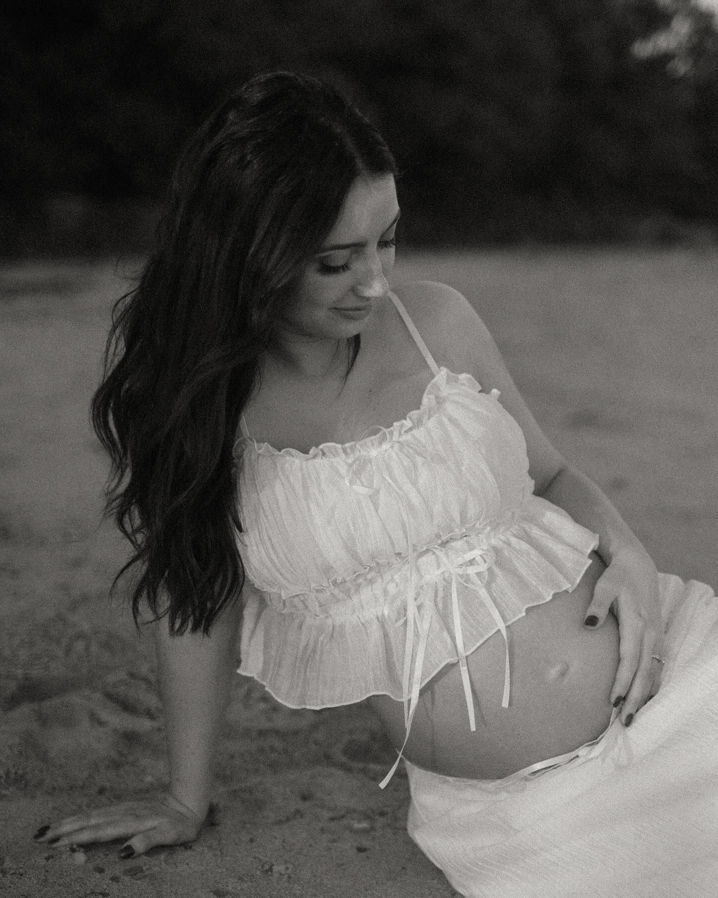 Black and white maternity photo of a pregnant woman in a white crop top sitting on sandy beach.