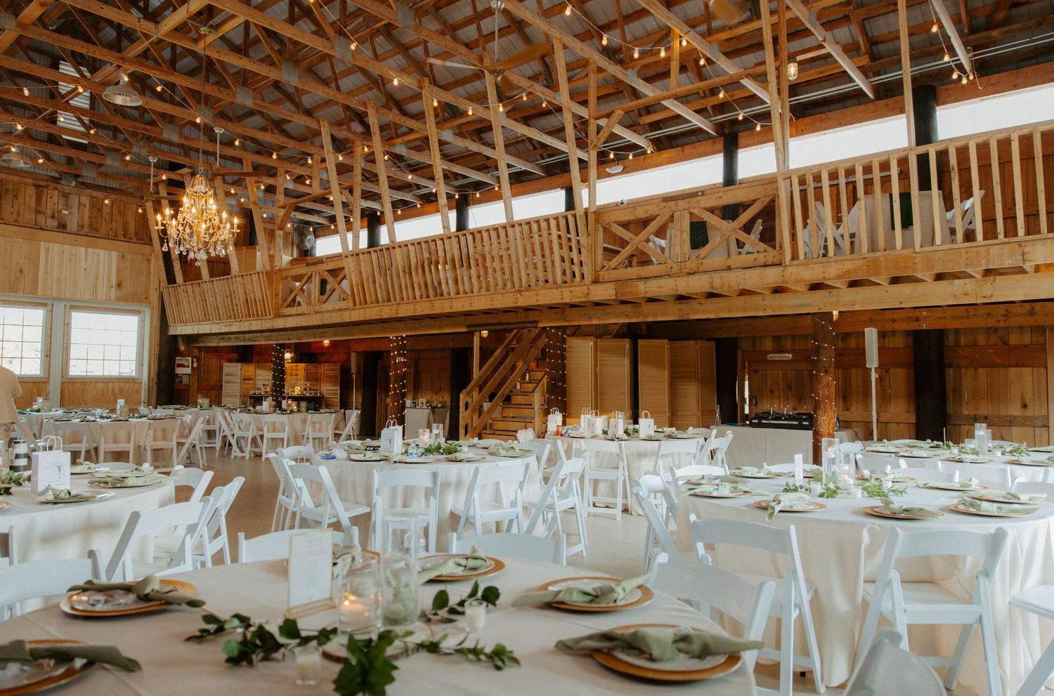Rustic barn wedding reception with white round tables, wooden chandeliers, and elegant greenery decor.