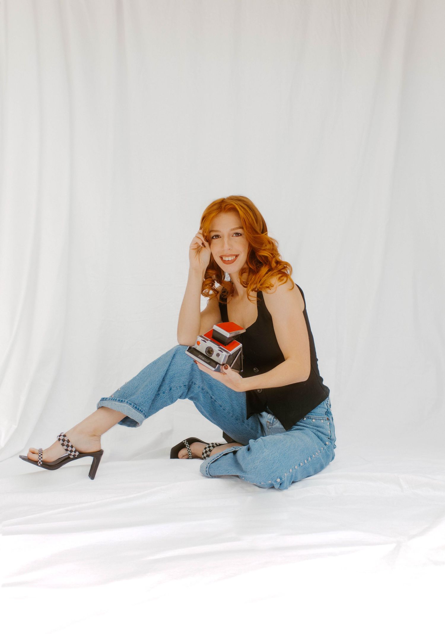 Red-haired woman in black tank and cuffed jeans sits on white backdrop holding a Polaroid camera.