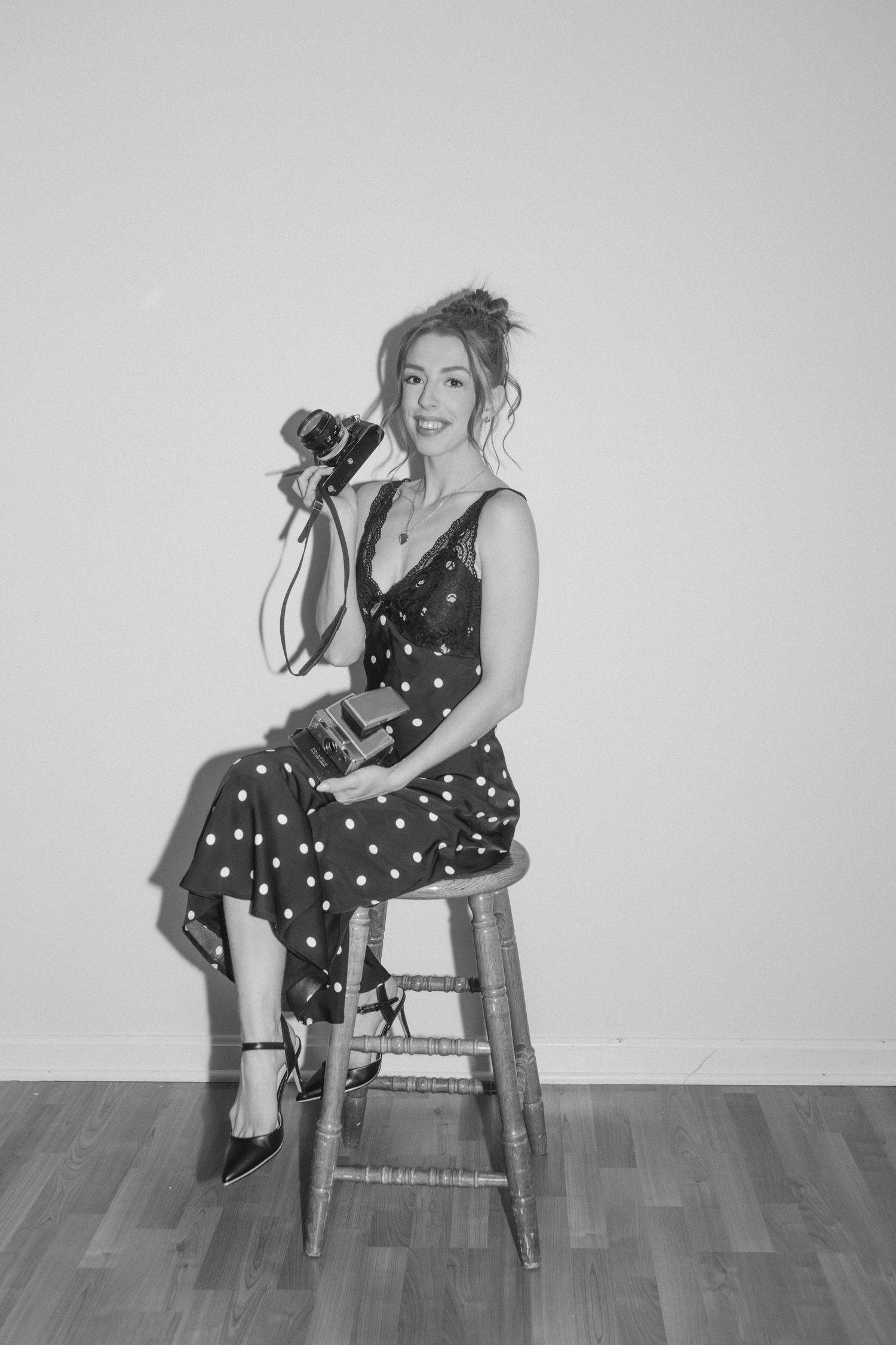 Woman in polka dot dress sitting on wooden stool holding vintage camera in black and white portrait.