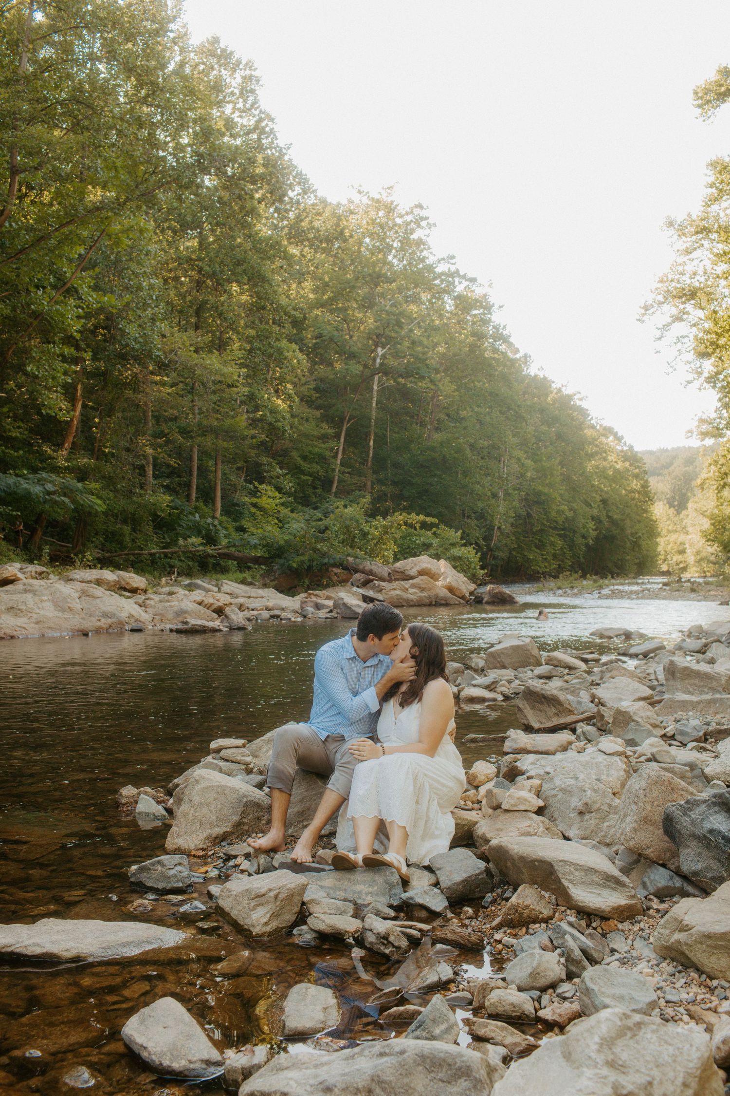 Couple sitting on rocks by a sunlit forest river, sharing a tender forehead touch during an engagement session.