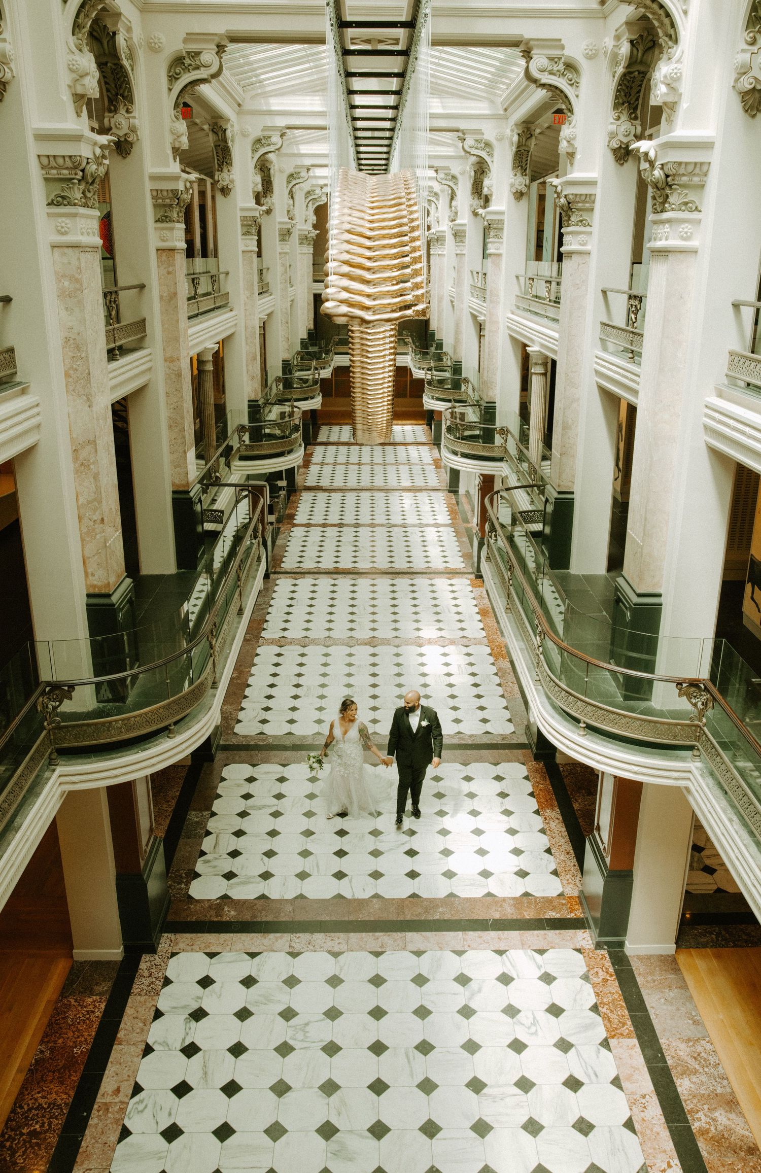 Bride and groom walking through elegant multi-story atrium with marble floors and ornate architecture at wedding venue.
