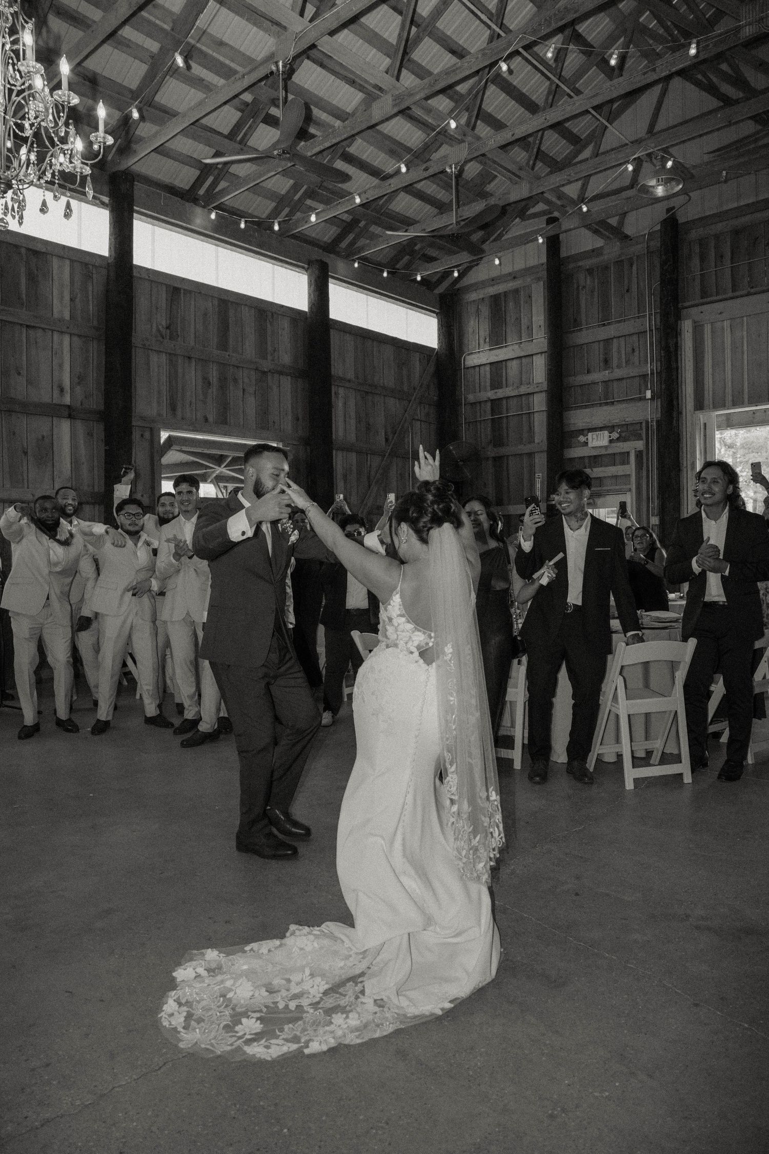 Black and white photo of bride and groom dancing surrounded by cheering wedding guests in barn venue.