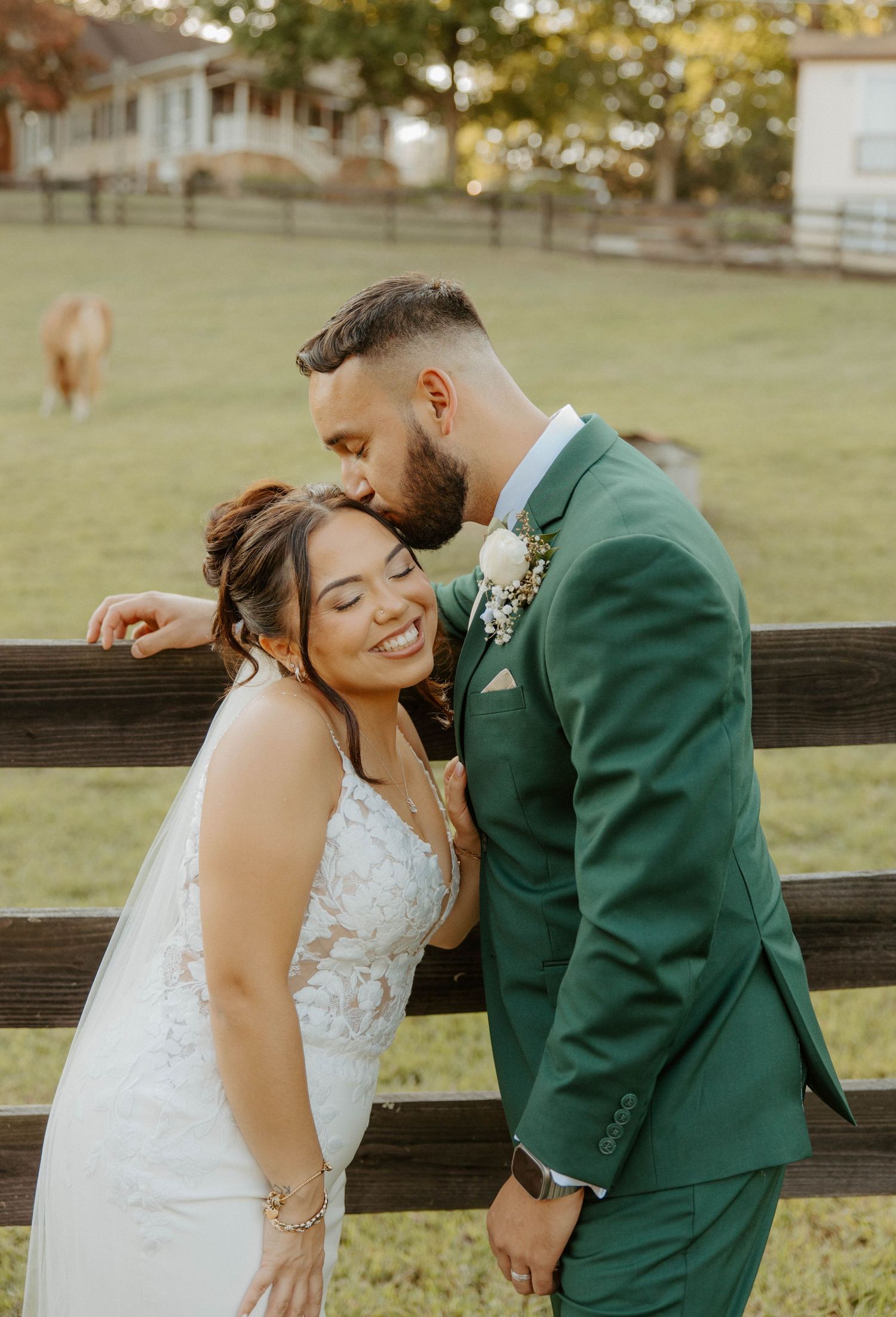 Smiling bride and groom in green suit posing by fence at farm wedding venue.