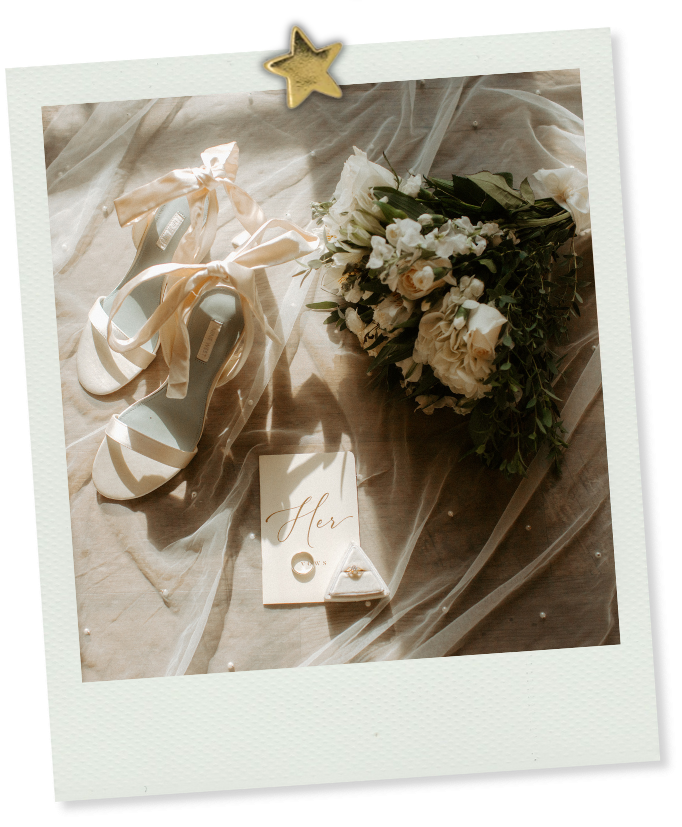 Elegant bridal flat lay with strappy heels, floral bouquet, rings, and a Her card on soft tulle fabric.