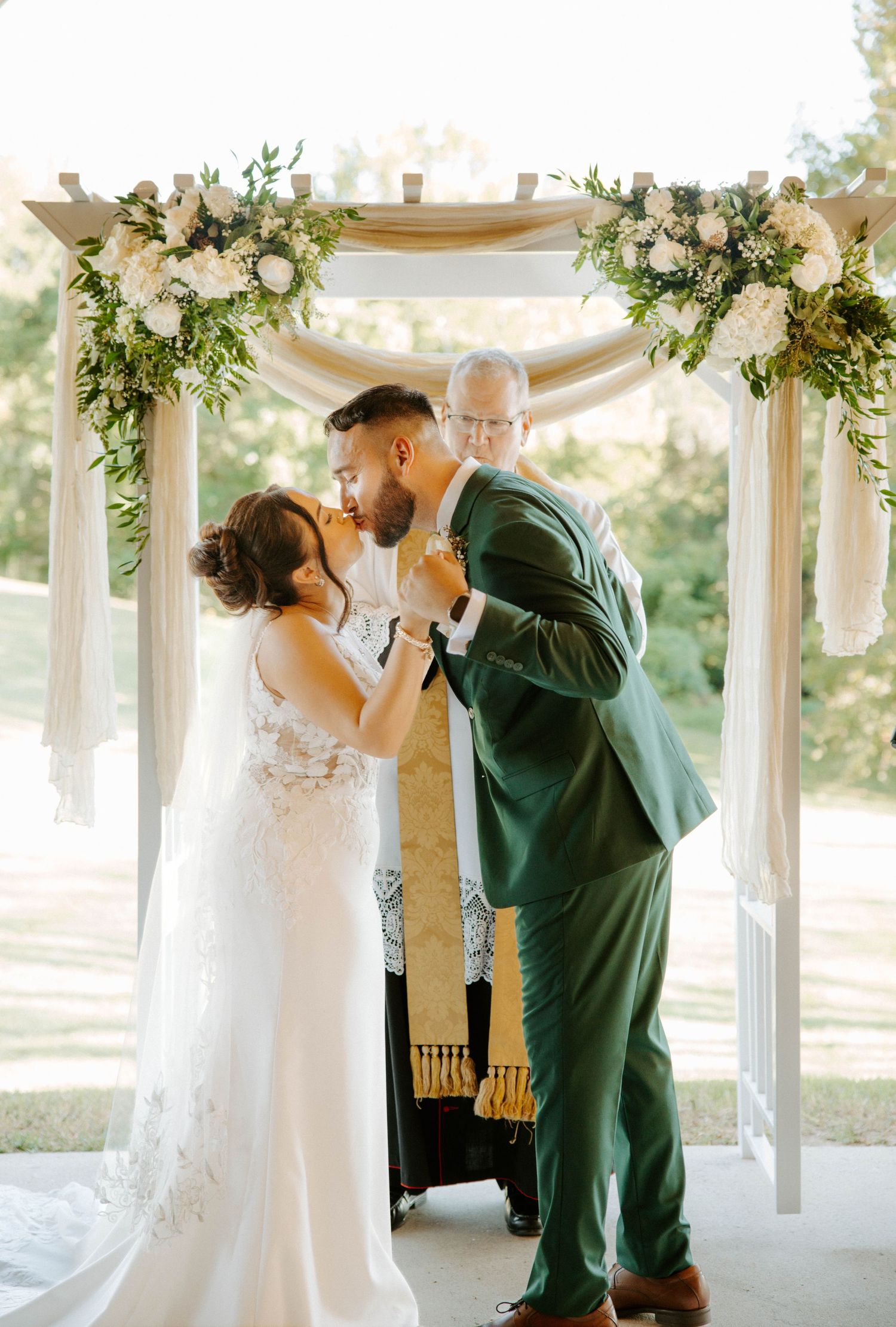Bride and groom sharing first kiss under floral greenery arch with officiant at ceremony.