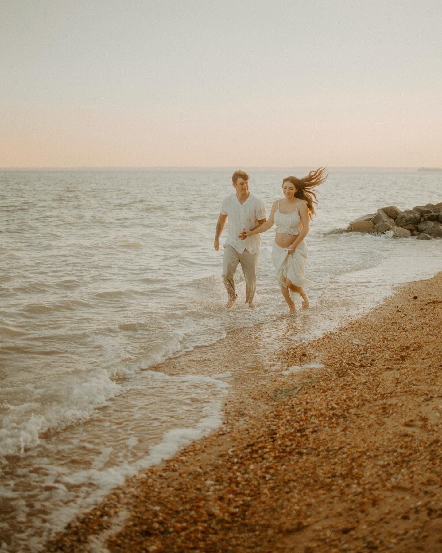 Joyful pregnant couple in white outfits running along a sandy beach shore at sunset.