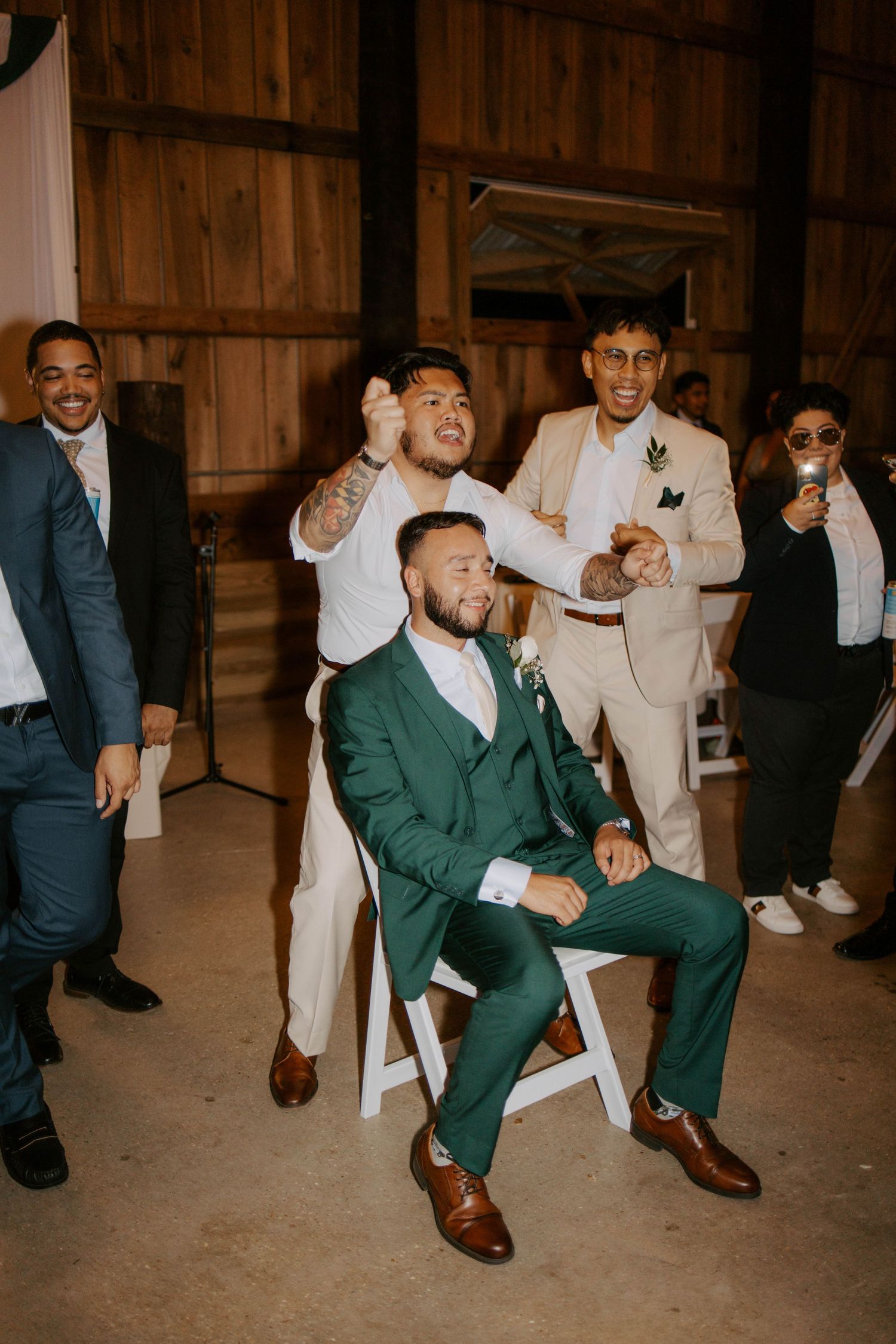Groom in green suit seated while groomsmen celebrate around him at barn reception.