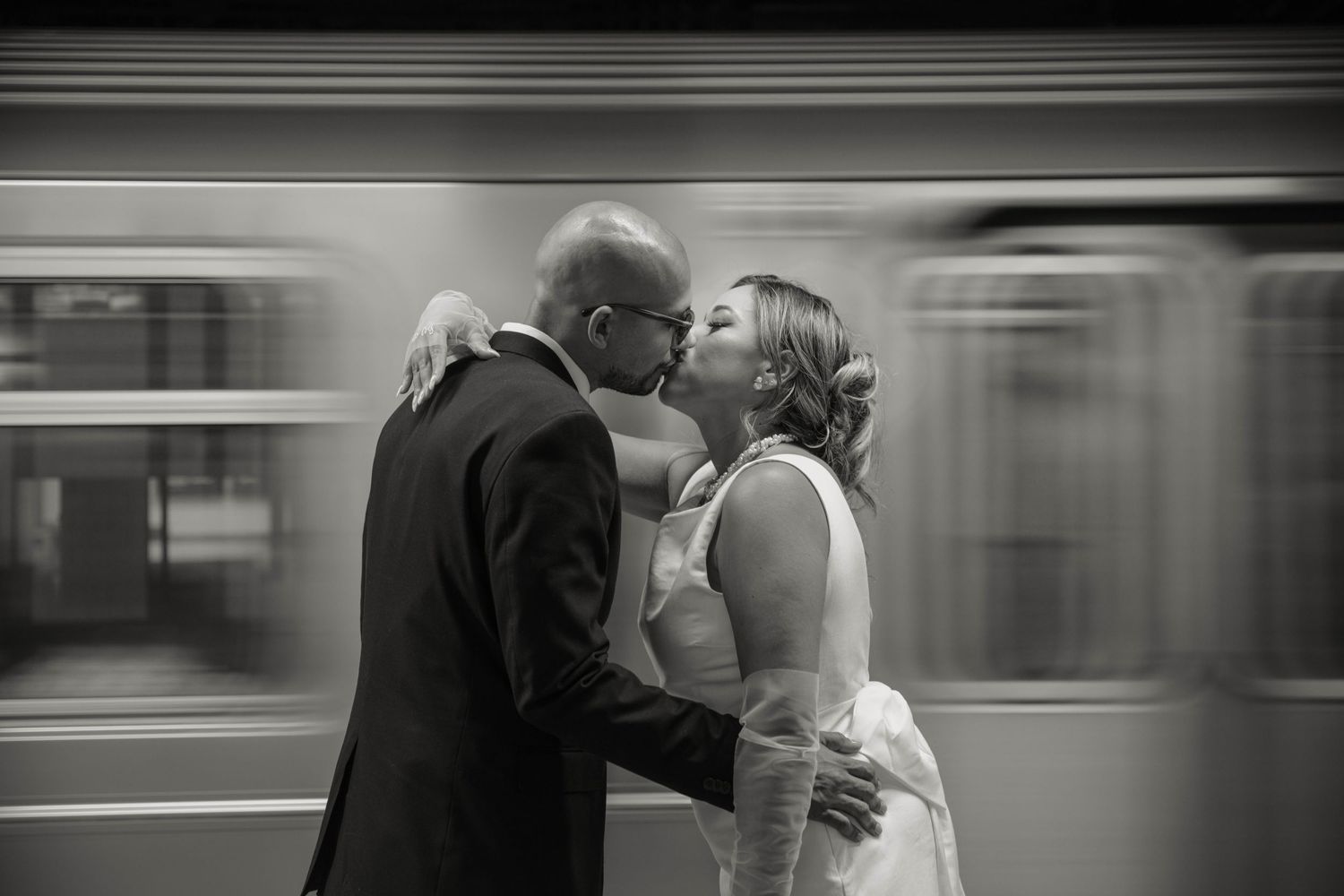 Black and white photo of bride and groom kissing on a subway platform as a train blurs past behind them.