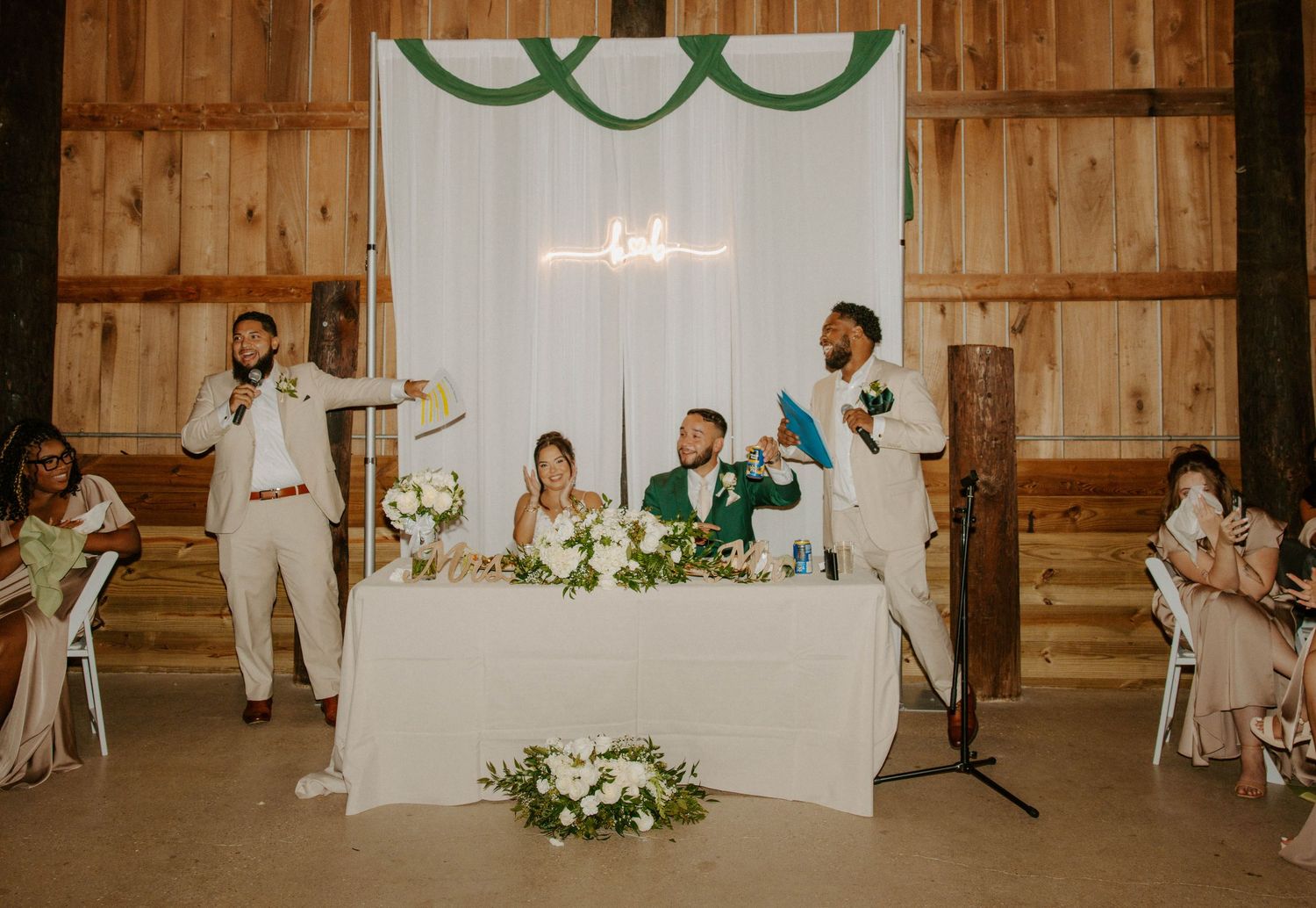 Groomsmen give toasts at rustic barn wedding reception with green and white sweetheart table.