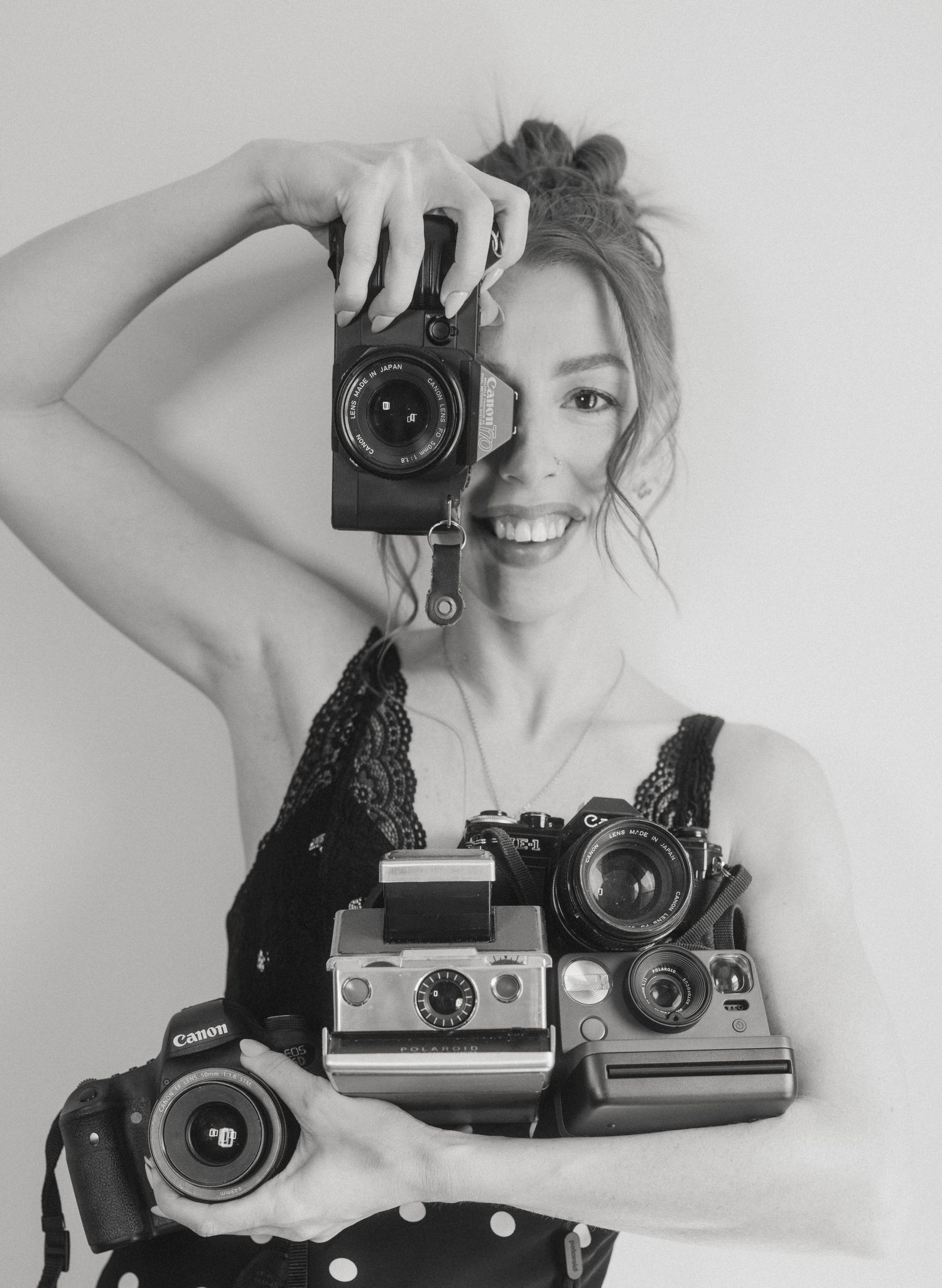 Smiling woman holding multiple vintage cameras including Canon and Polaroid in black and white photo.