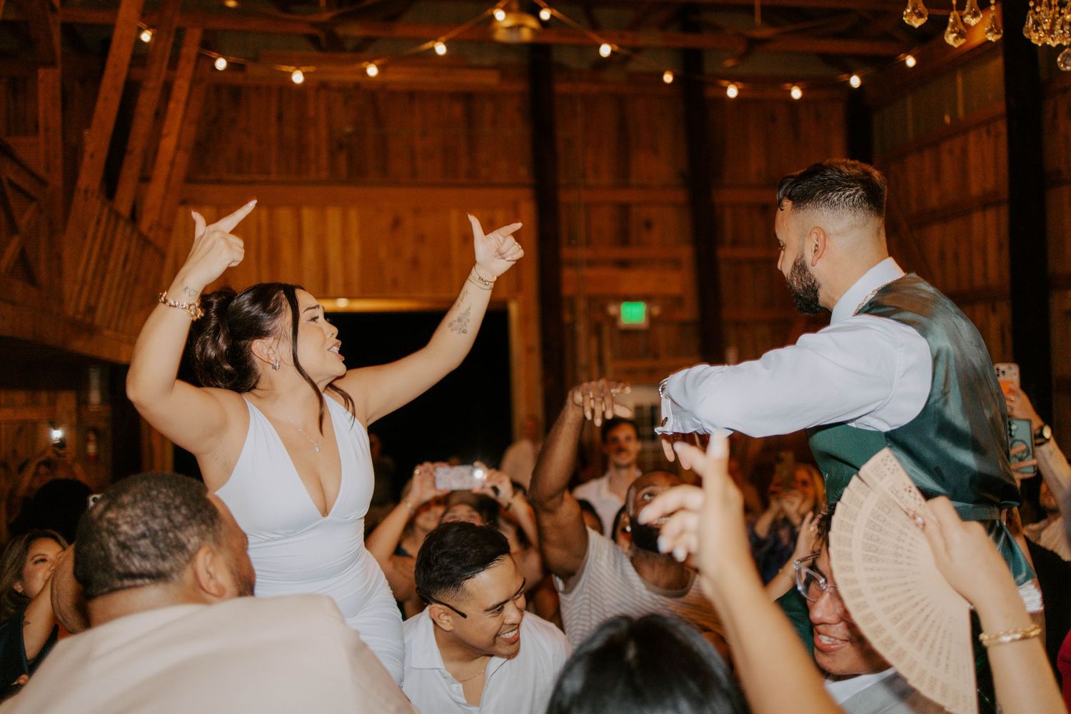 Bride and groom dancing with guests under string lights at rustic barn reception.