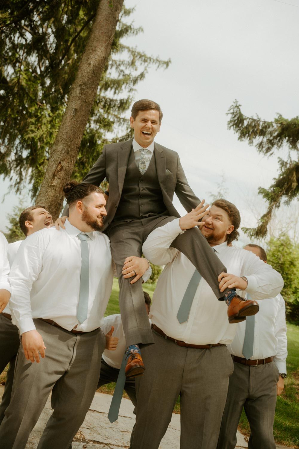 Laughing groom lifted by groomsmen in white shirts and gray pants during fun outdoor wedding celebration.