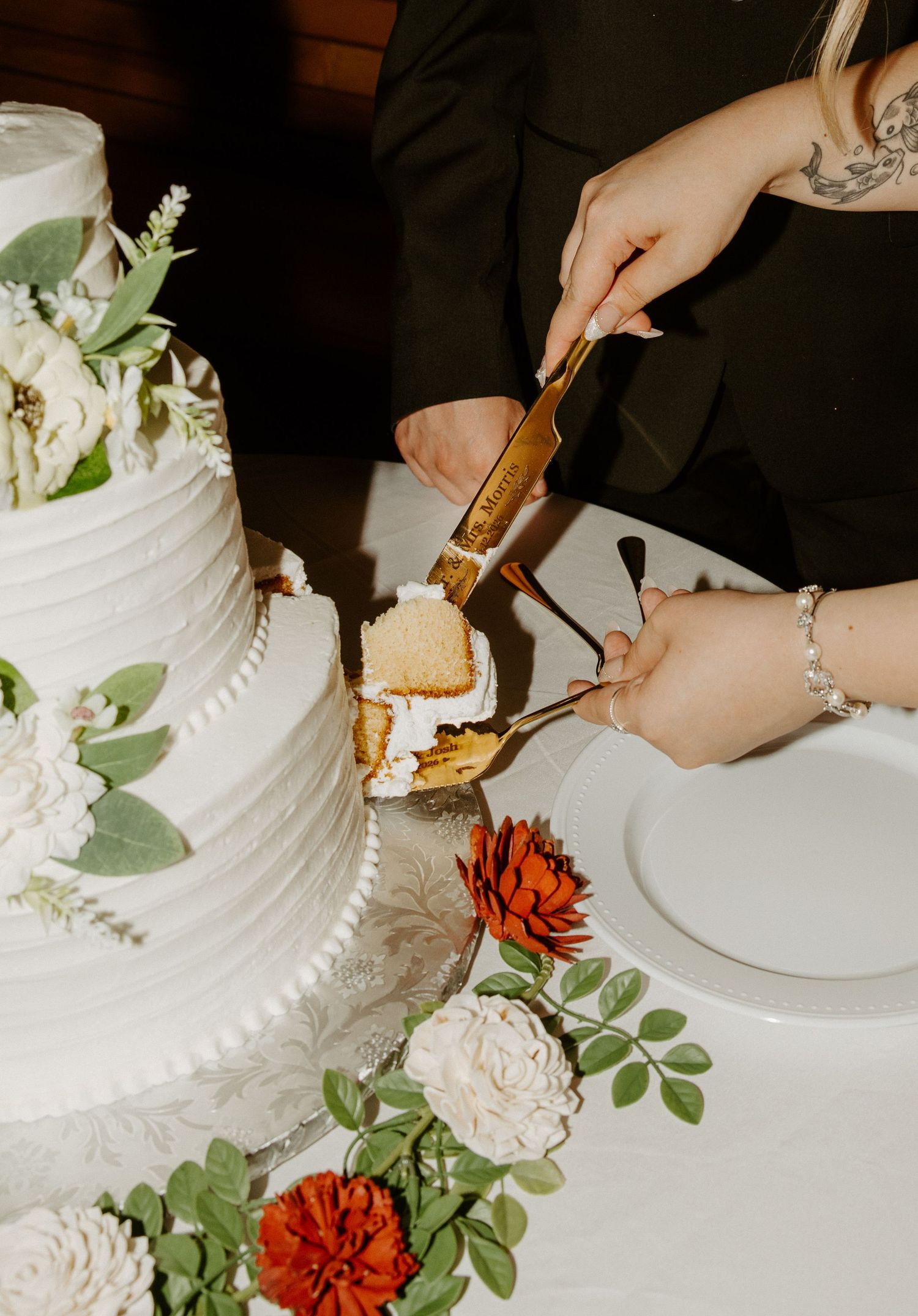Couple cutting a white tiered wedding cake adorned with white flowers and greenery using gold cake servers.