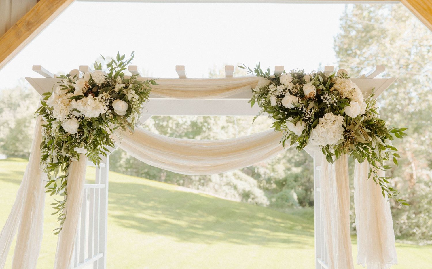 Elegant wedding arch adorned with white roses, greenery, and flowing white fabric draping outdoors.