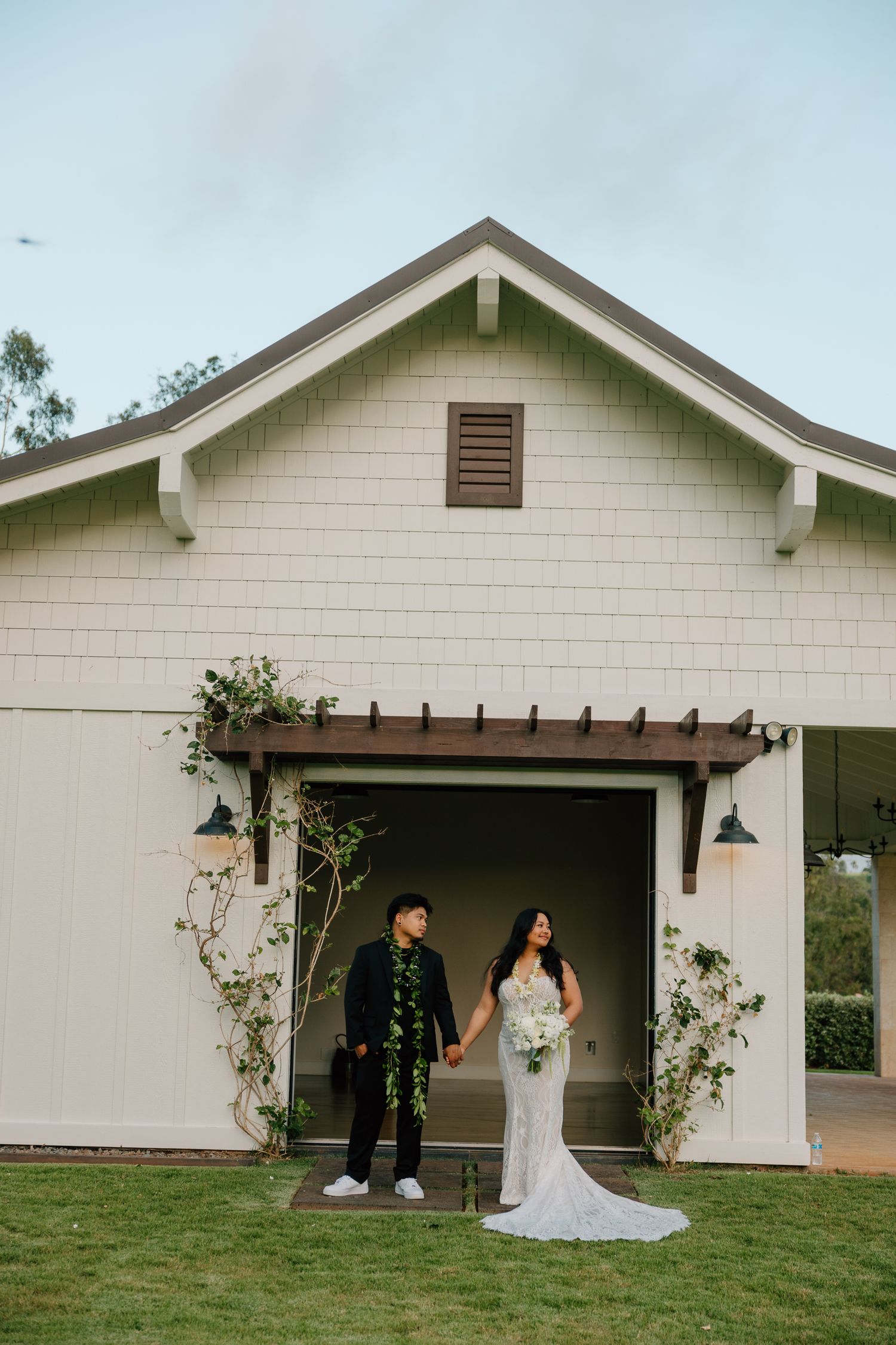 Bride and groom hold hands at barn entrance adorned with greenery at outdoor wedding venue.