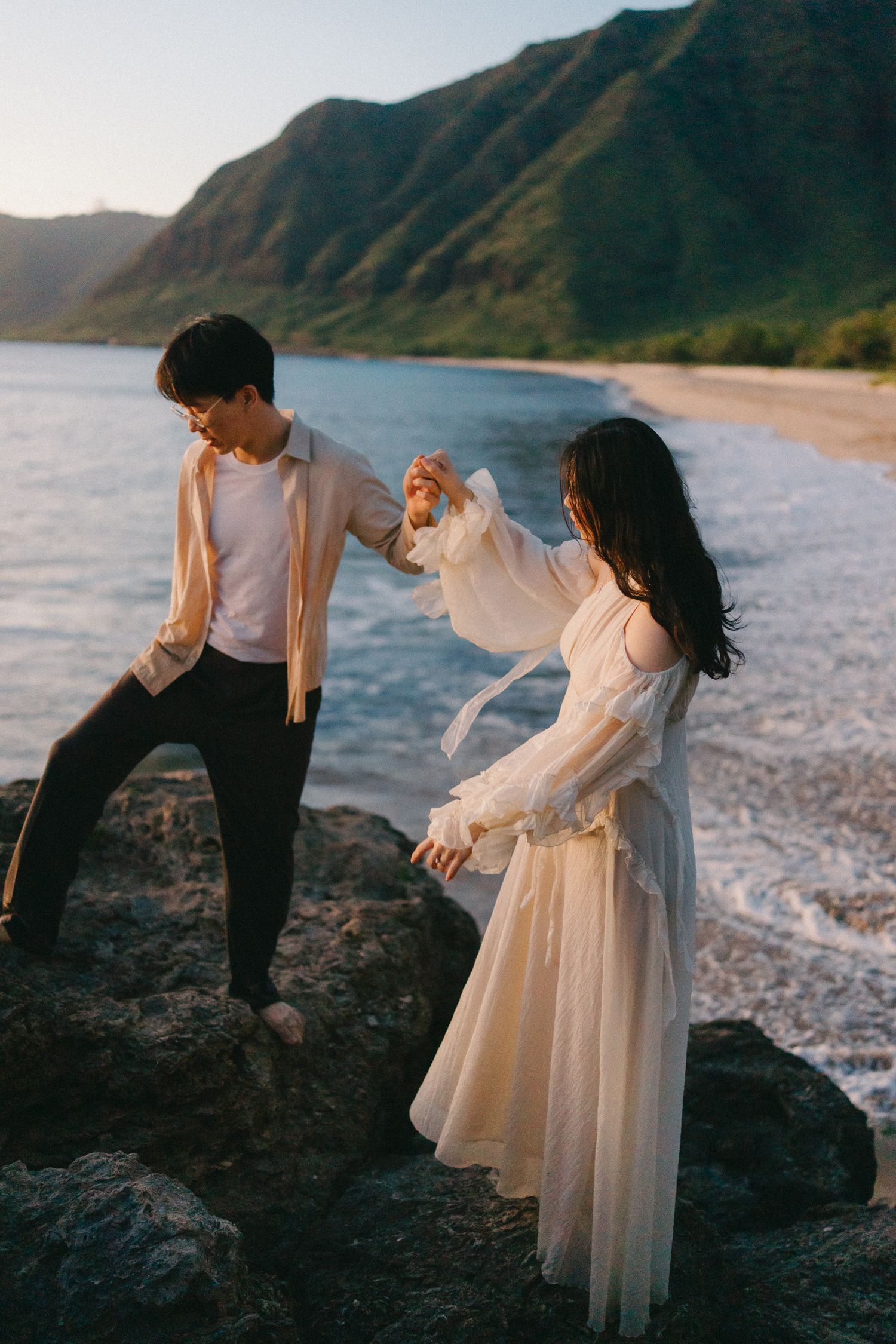 Couple holding hands on rocky beach shore with green mountains and ocean waves in background at sunset.