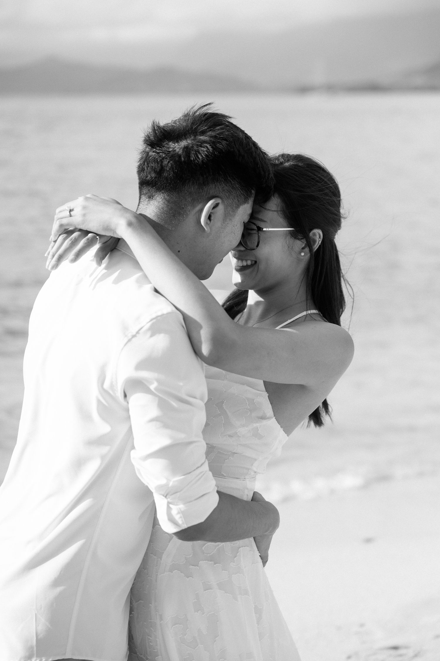 Black and white photo of a couple embracing and smiling at each other on a beach.