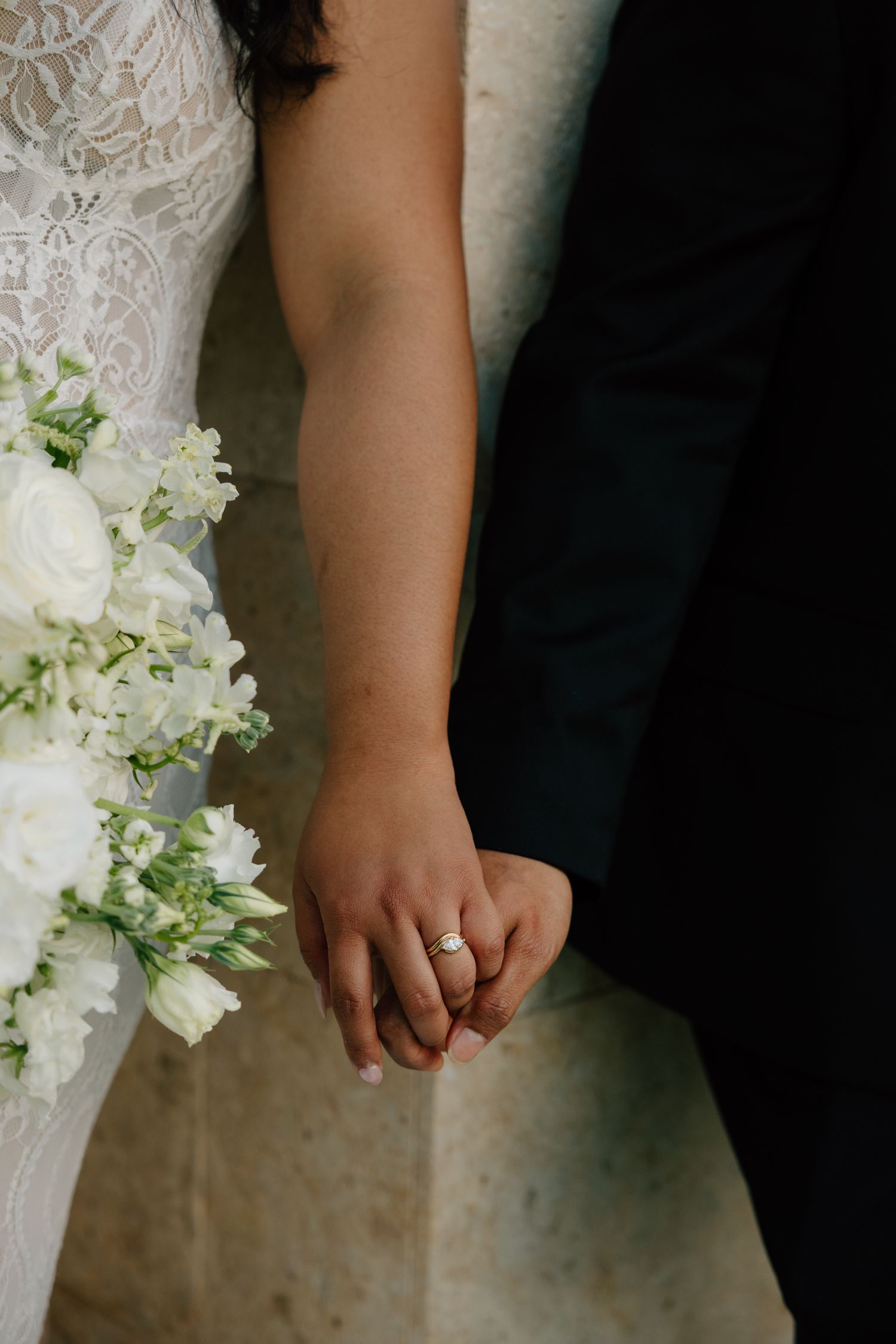 Bride and groom holding hands, showcasing an elegant engagement ring, with a white floral bouquet at a wedding ceremony.