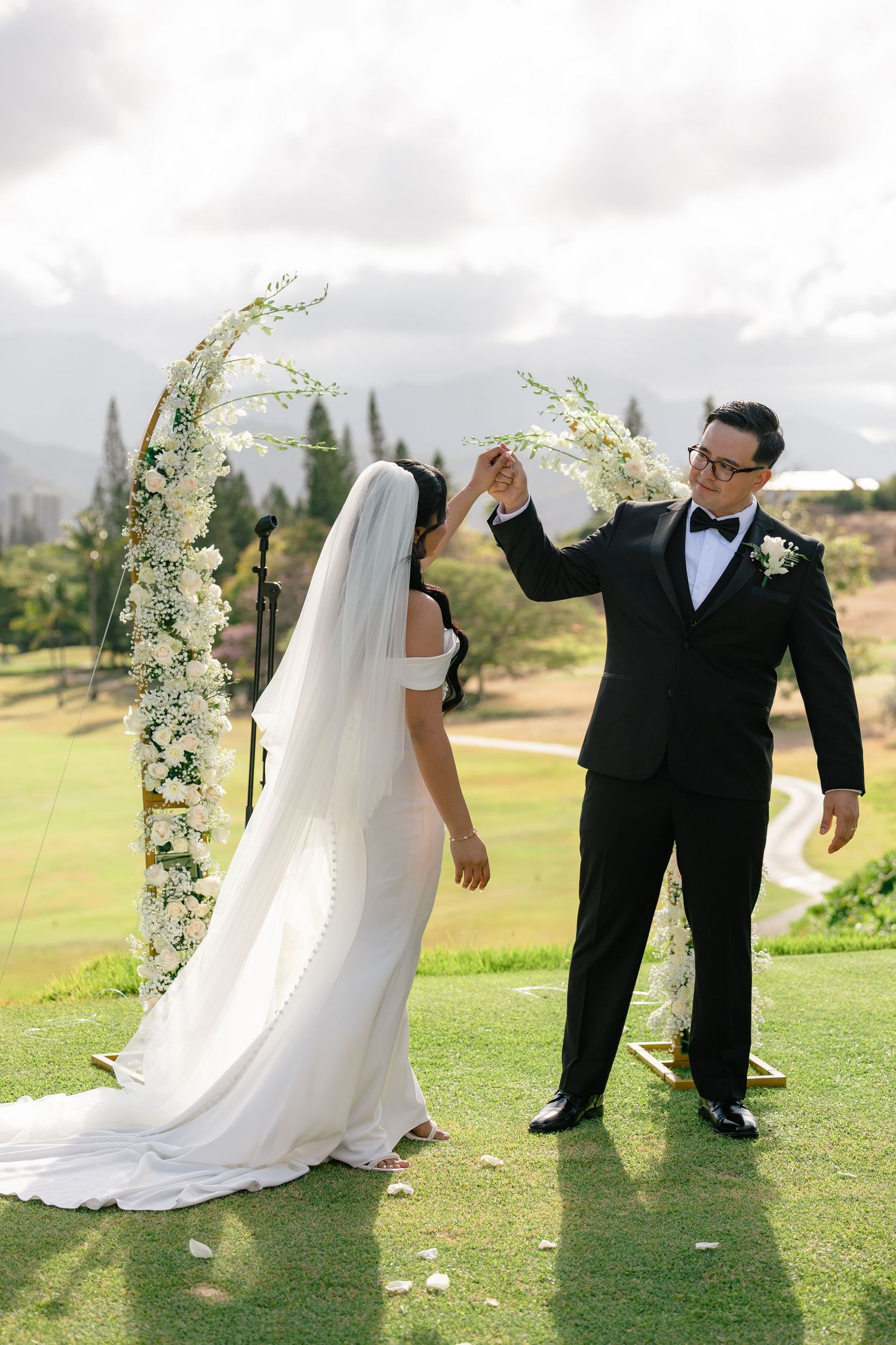 Groom in black tuxedo spinning bride in white gown under floral arch on golf course during outdoor wedding ceremony.