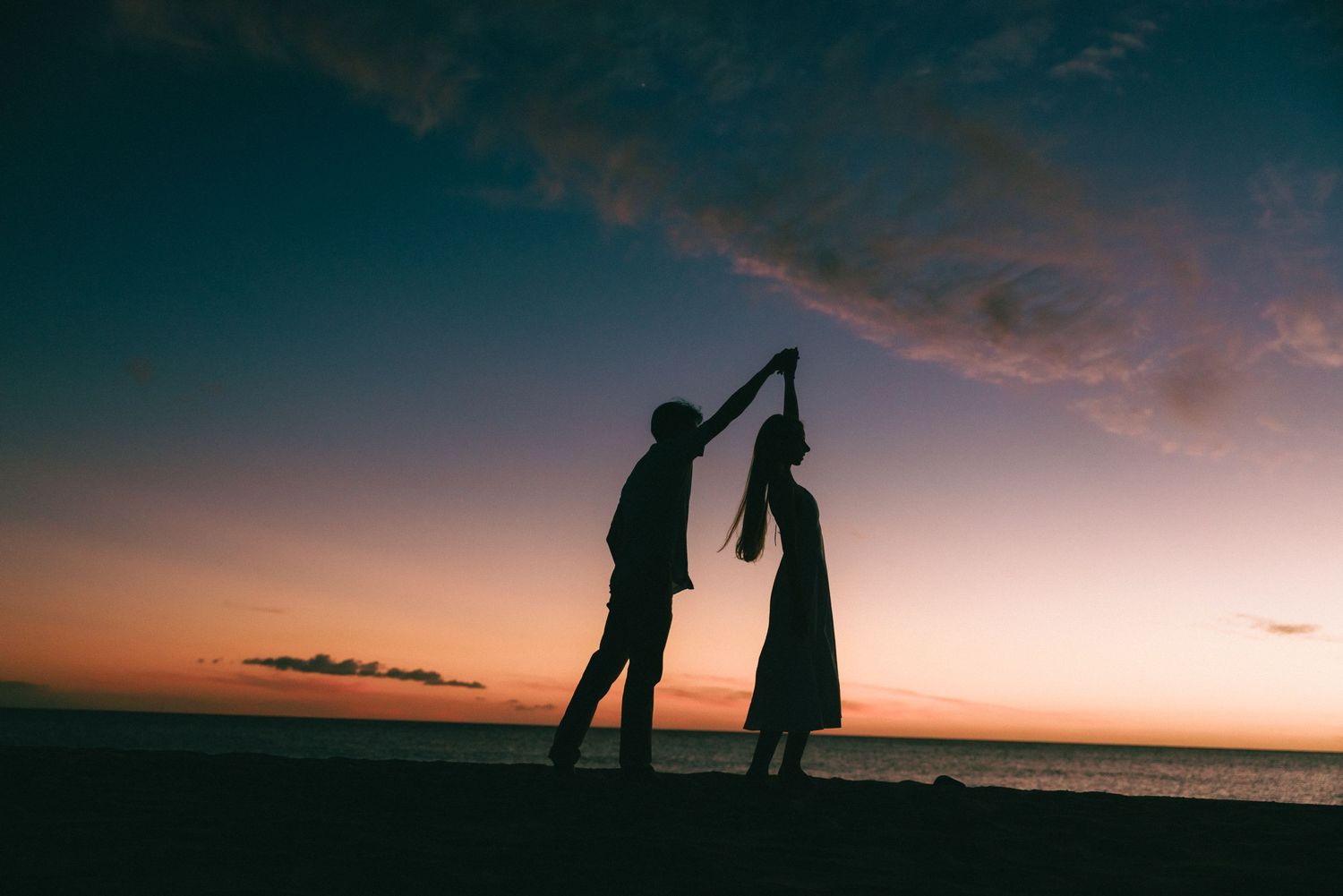 Silhouette of a couple dancing on a beach at sunset, with a colorful sky and calm ocean in the background.