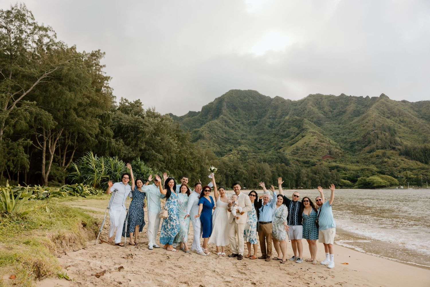 A wedding party group together on a beach with the bride and groom -- lush Hawaiian mountain backdrop with warm sandy beach.
