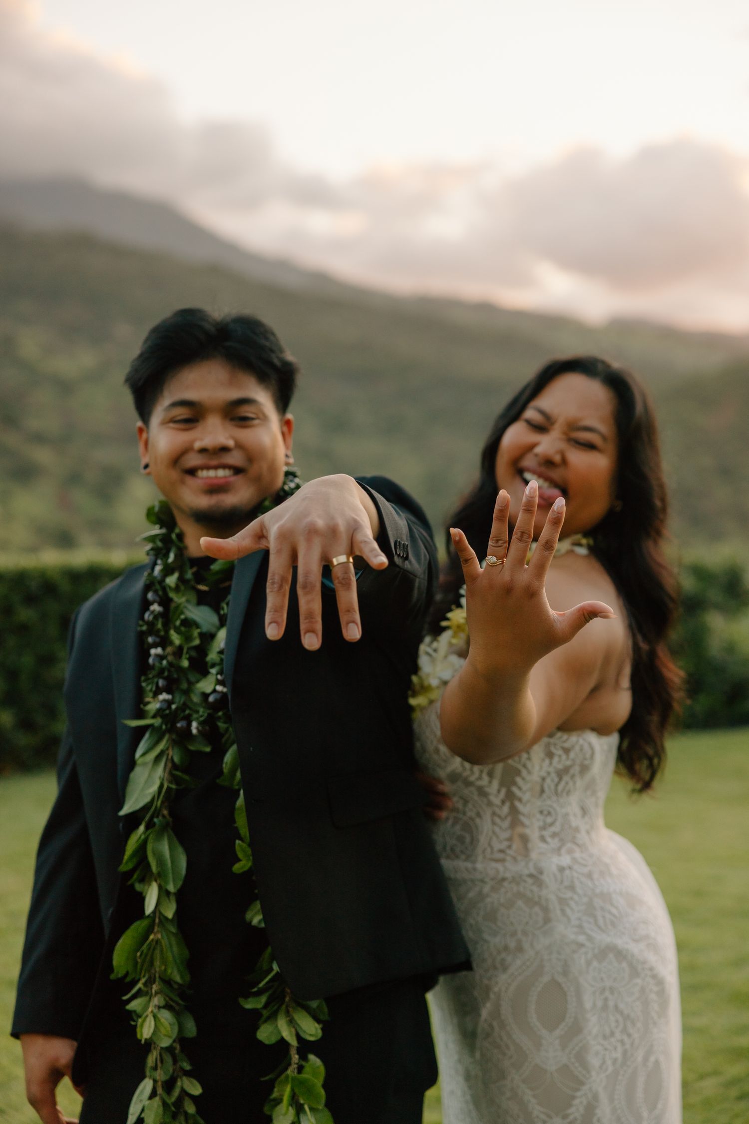 Newlywed couple in Hawaii showing off wedding rings, groom in black suit with lei, bride in lace gown, mountains behind.