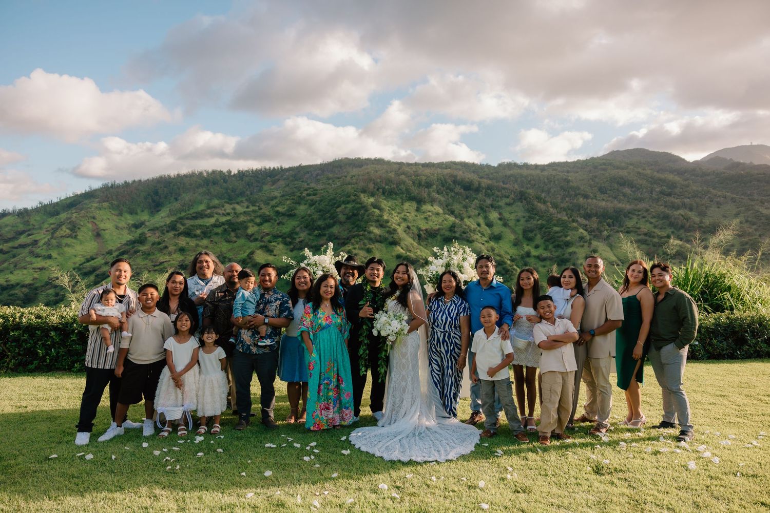 Wedding group photo with bride and groom surrounded by family on green lawn with lush mountain backdrop.