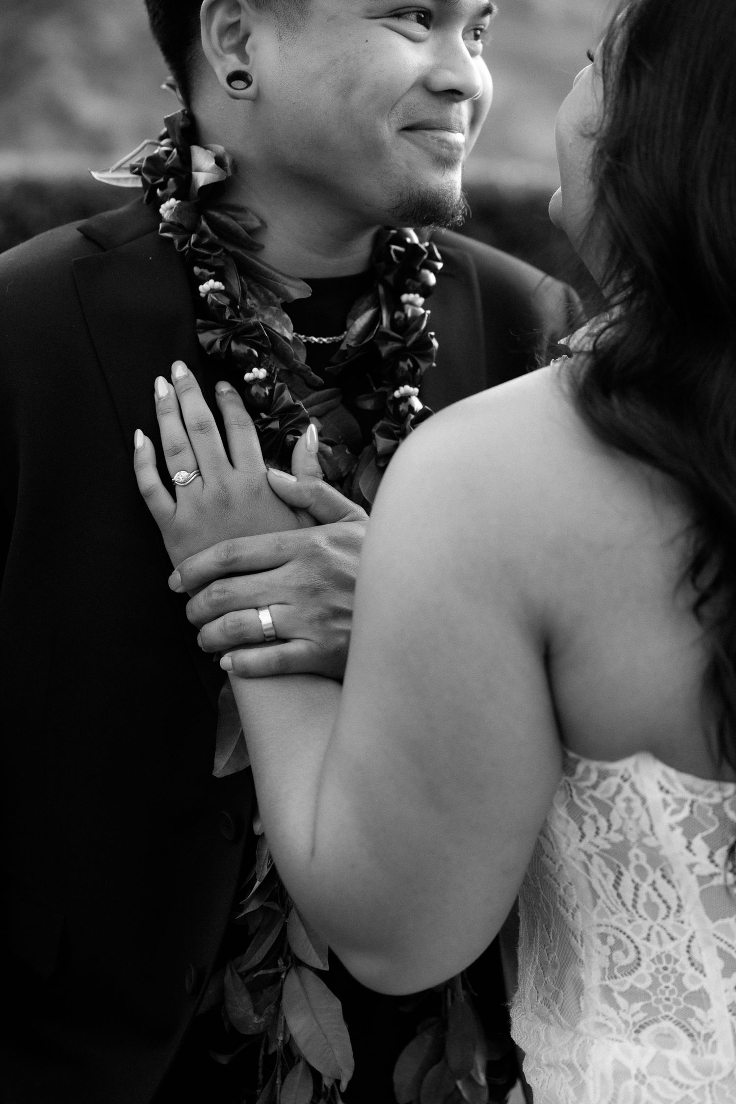 Close-up black and white wedding photo showing bride and groom's hands with rings, groom wearing a tropical lei.