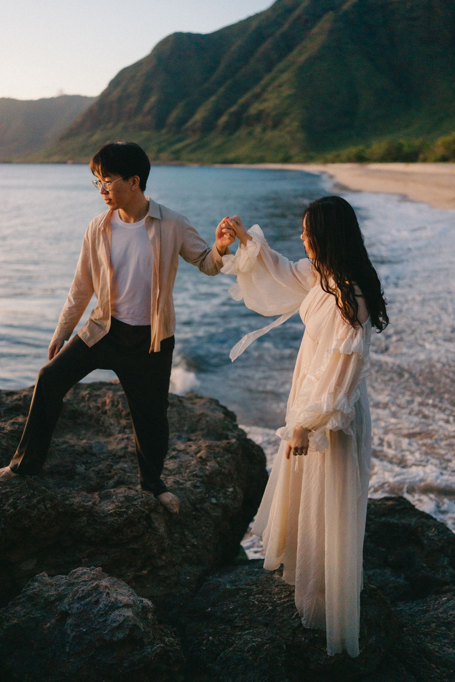 Couple holding hands on rocky beach with ocean waves and green mountains in the background at sunset.