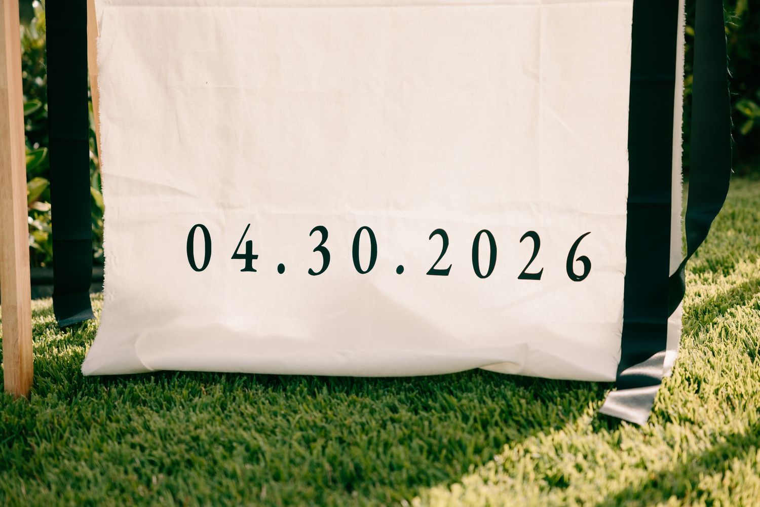 White fabric banner displaying wedding date 04.30.2026 in elegant black serif font on green grass.