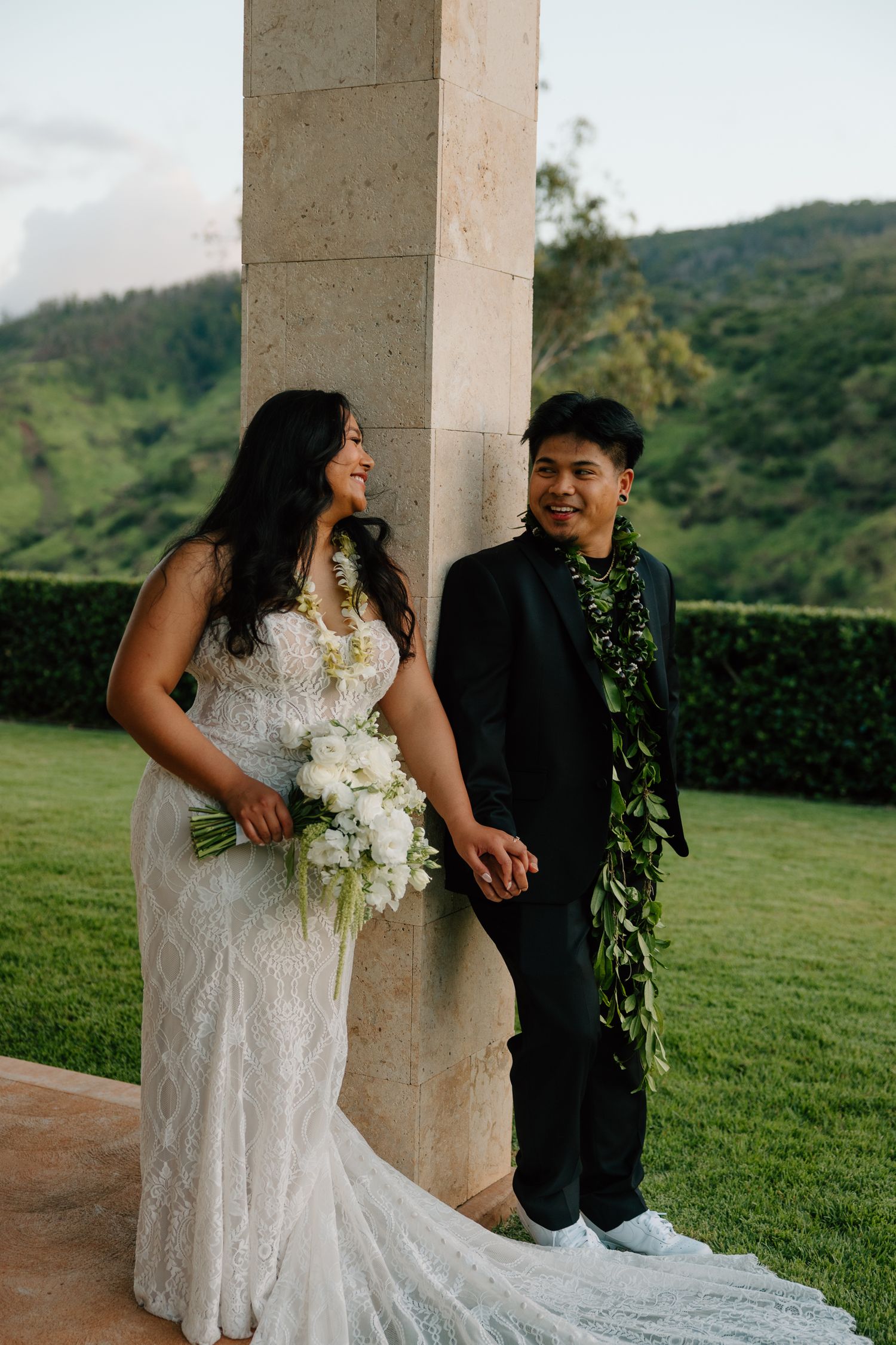 Bride in lace gown holding white bouquet stands with groom in black suit wearing green lei, scenic green hills backdrop.