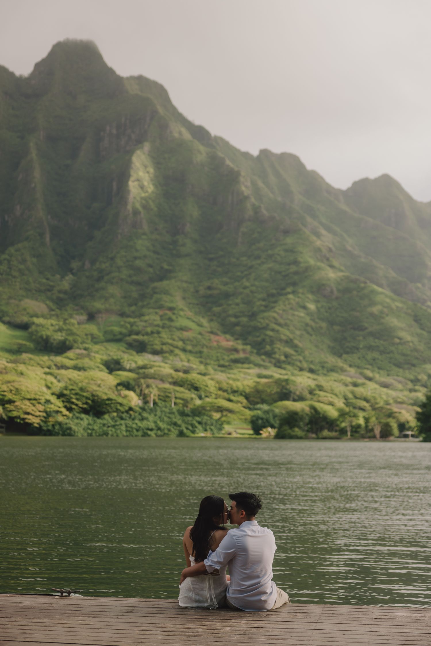 A couple sits on a dock sharing a kiss with lush Hawaiian mountains and calm water behind them.