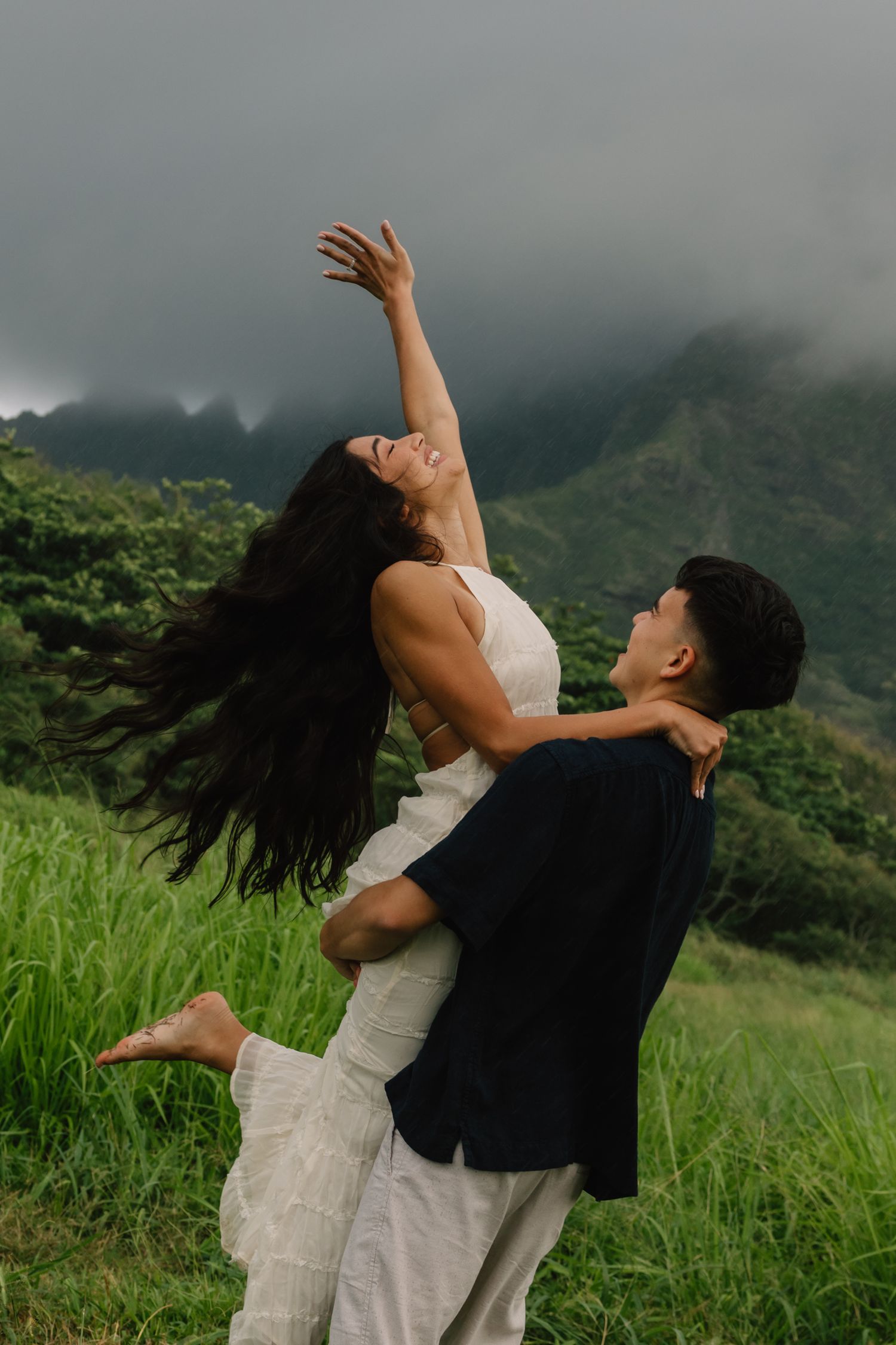 A couple dances joyfully in a lush green Hawaiian valley, woman arching back with arm raised, misty mountains behind.