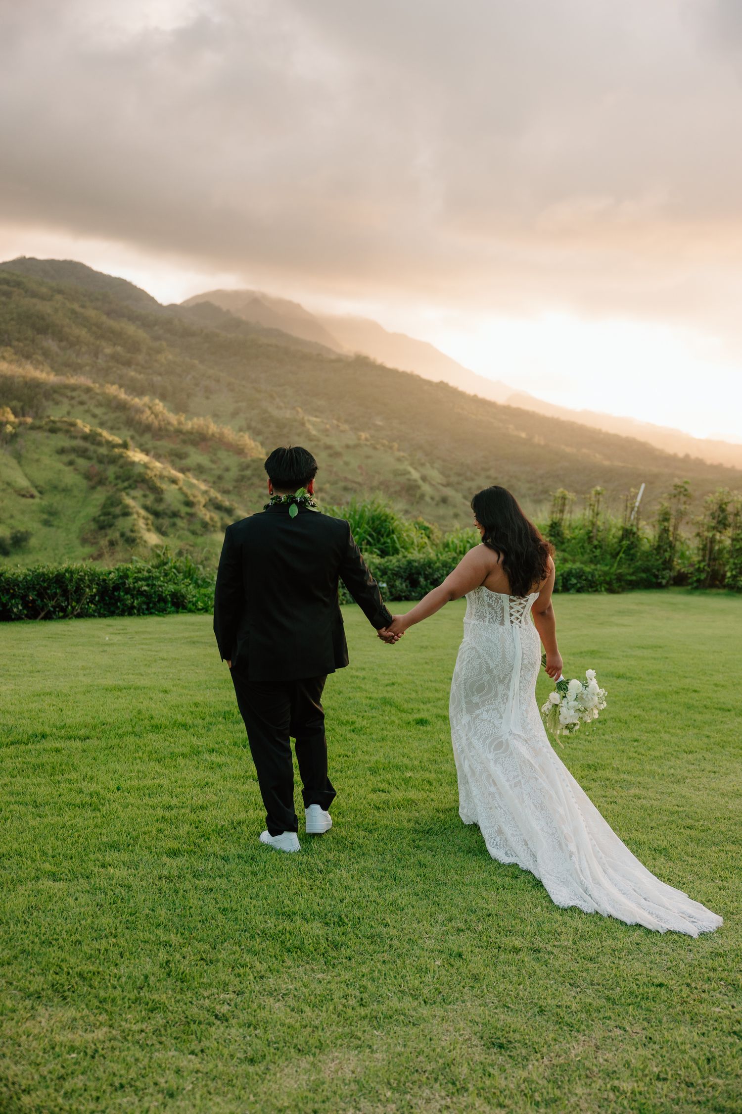 Bride and groom walk toward stunning Hawaii mountain sunset, bride in lace gown holding white bouquet, groom in black suit.