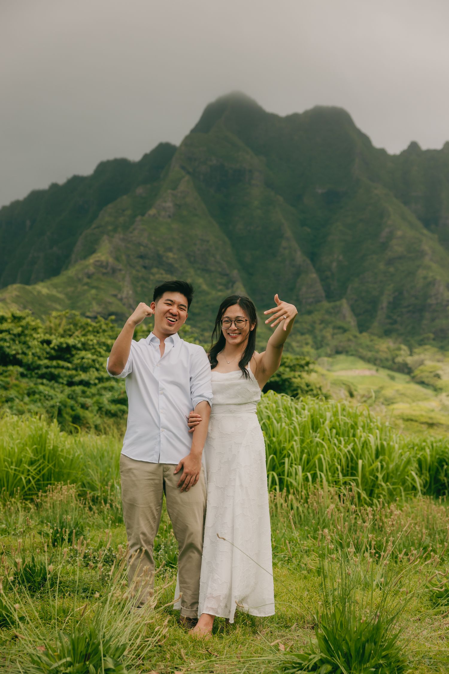 A joyful couple poses in front of lush green mountains in Hawaii, woman in white dress, man in light blue shirt.