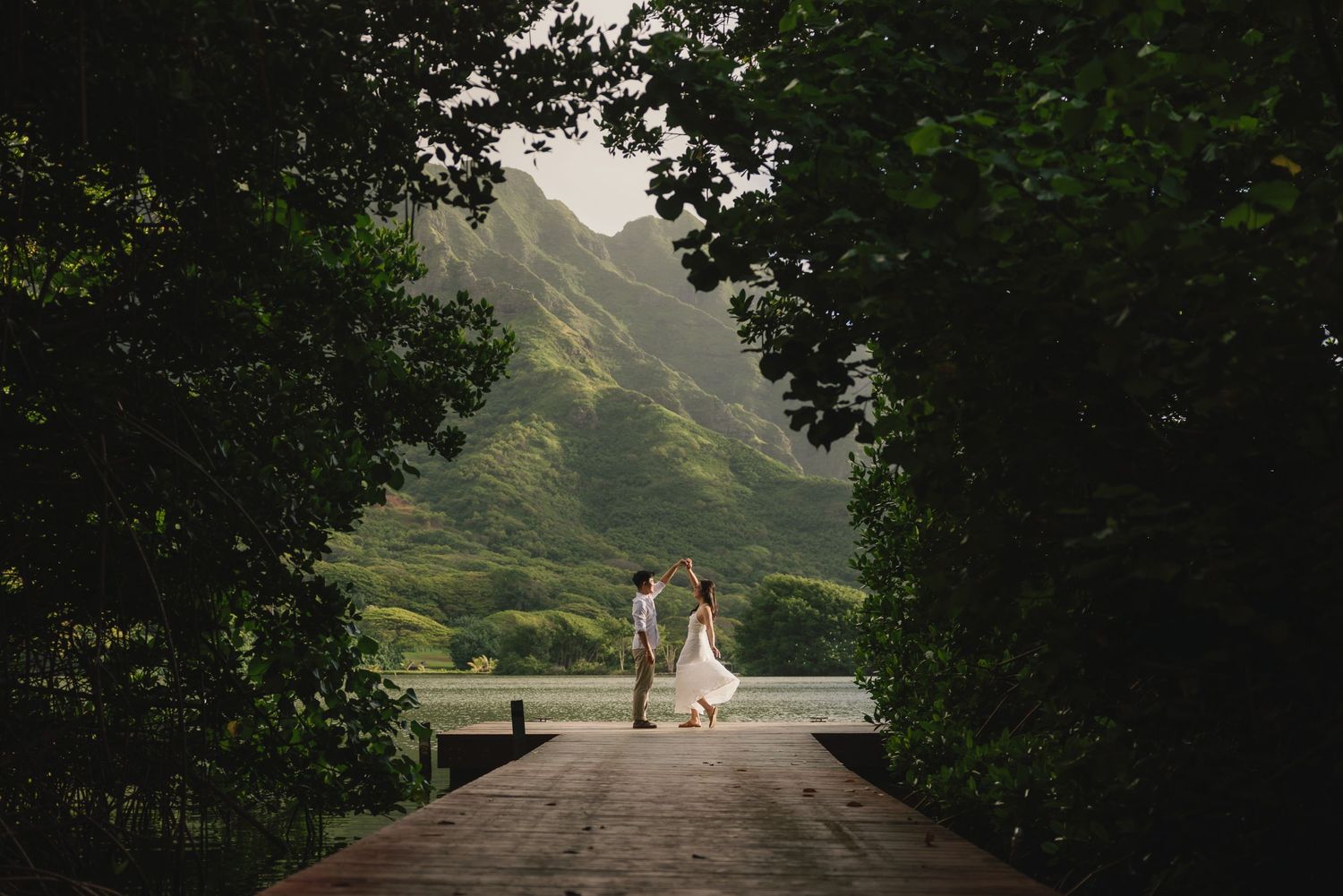 Couple walking hand in hand on a wooden path framed by lush green trees with misty mountains in Hawaii.