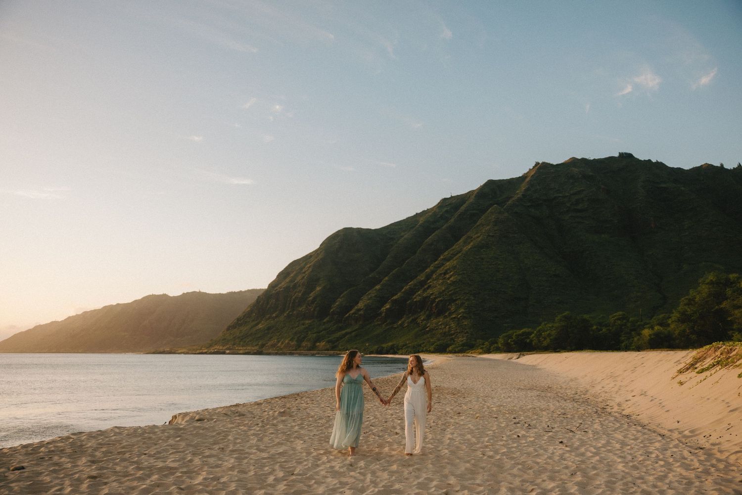 Two women hold hands on a sandy Hawaiian beach at sunset with lush green mountains in the background.