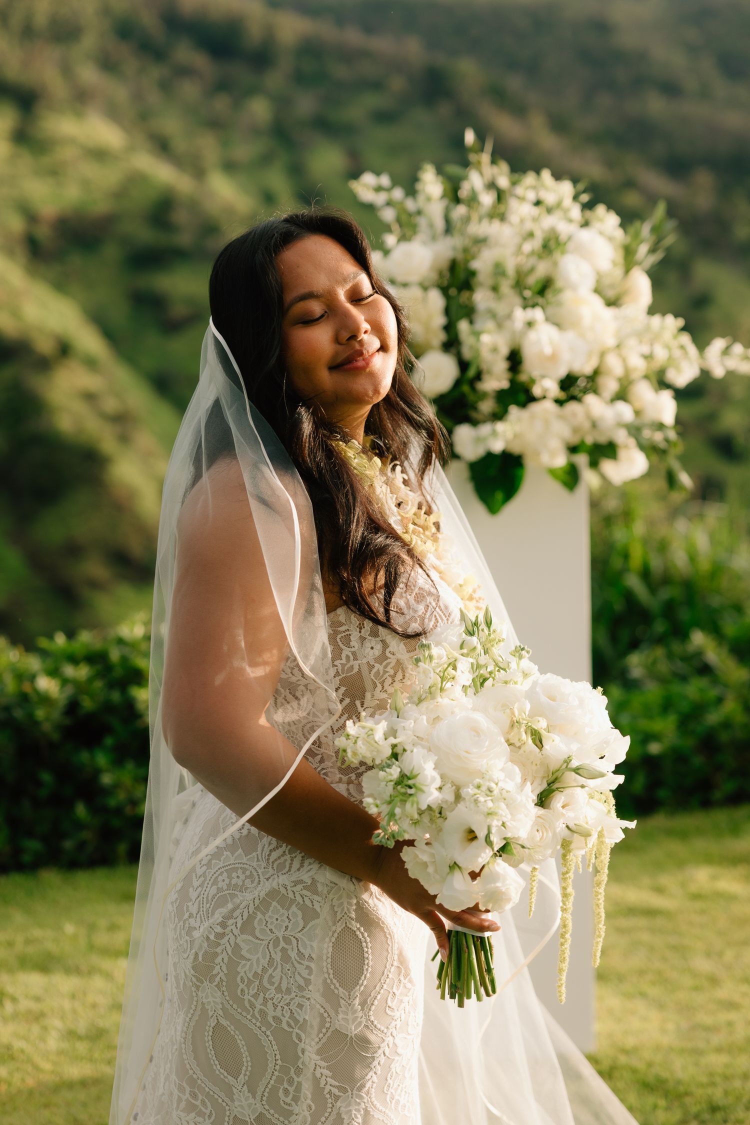 Smiling bride in lace gown and veil holds white bouquet outdoors with lush green hills backdrop.