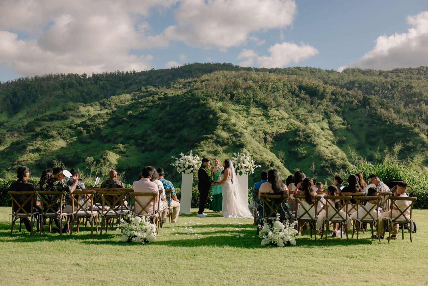 Outdoor wedding ceremony on lush green lawn with mountain backdrop, guests seated at rustic wooden tables in Hawaii.
