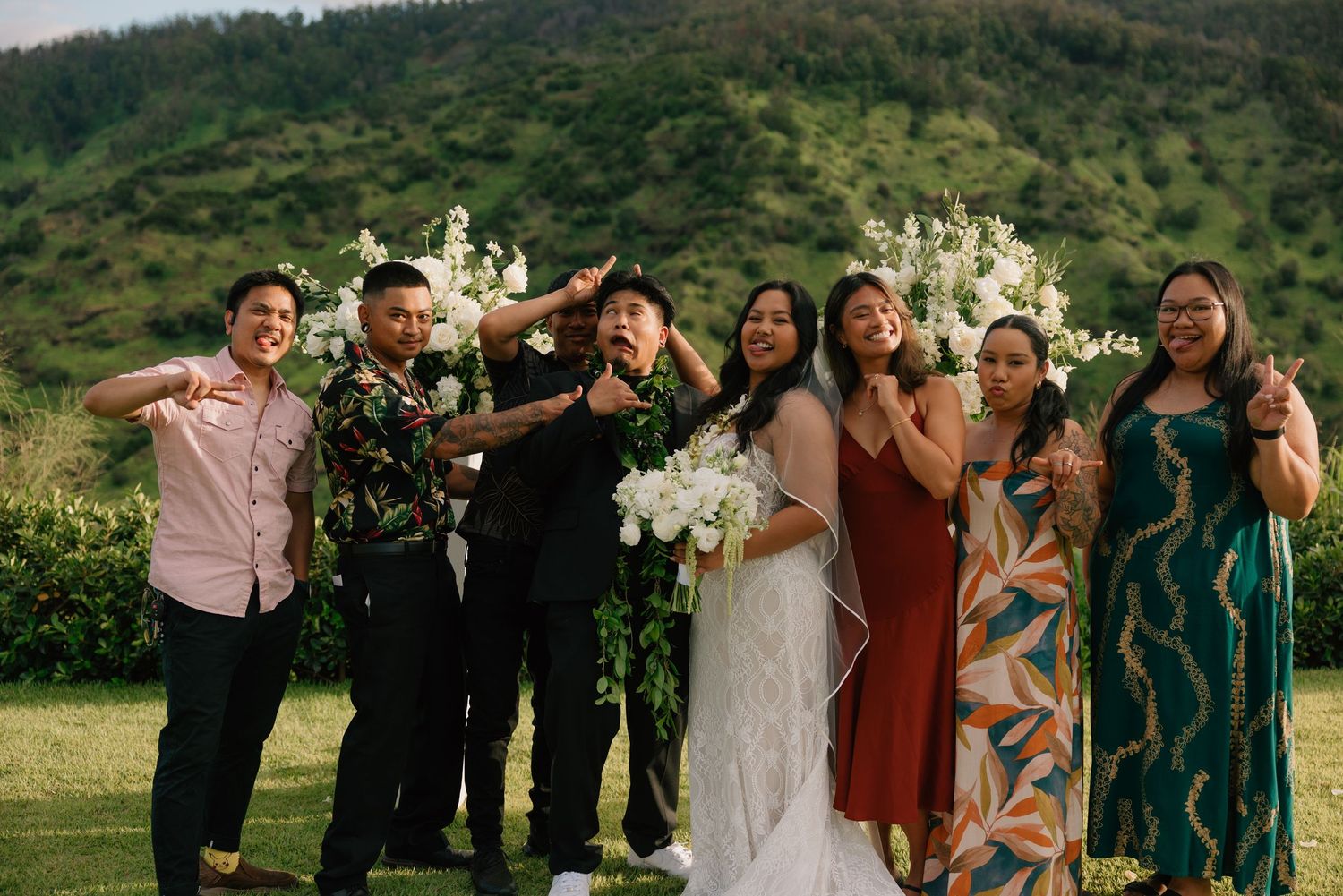 Wedding party posing joyfully outdoors in Hawaii with lush green mountains, bride holding white bouquet in elegant gown.