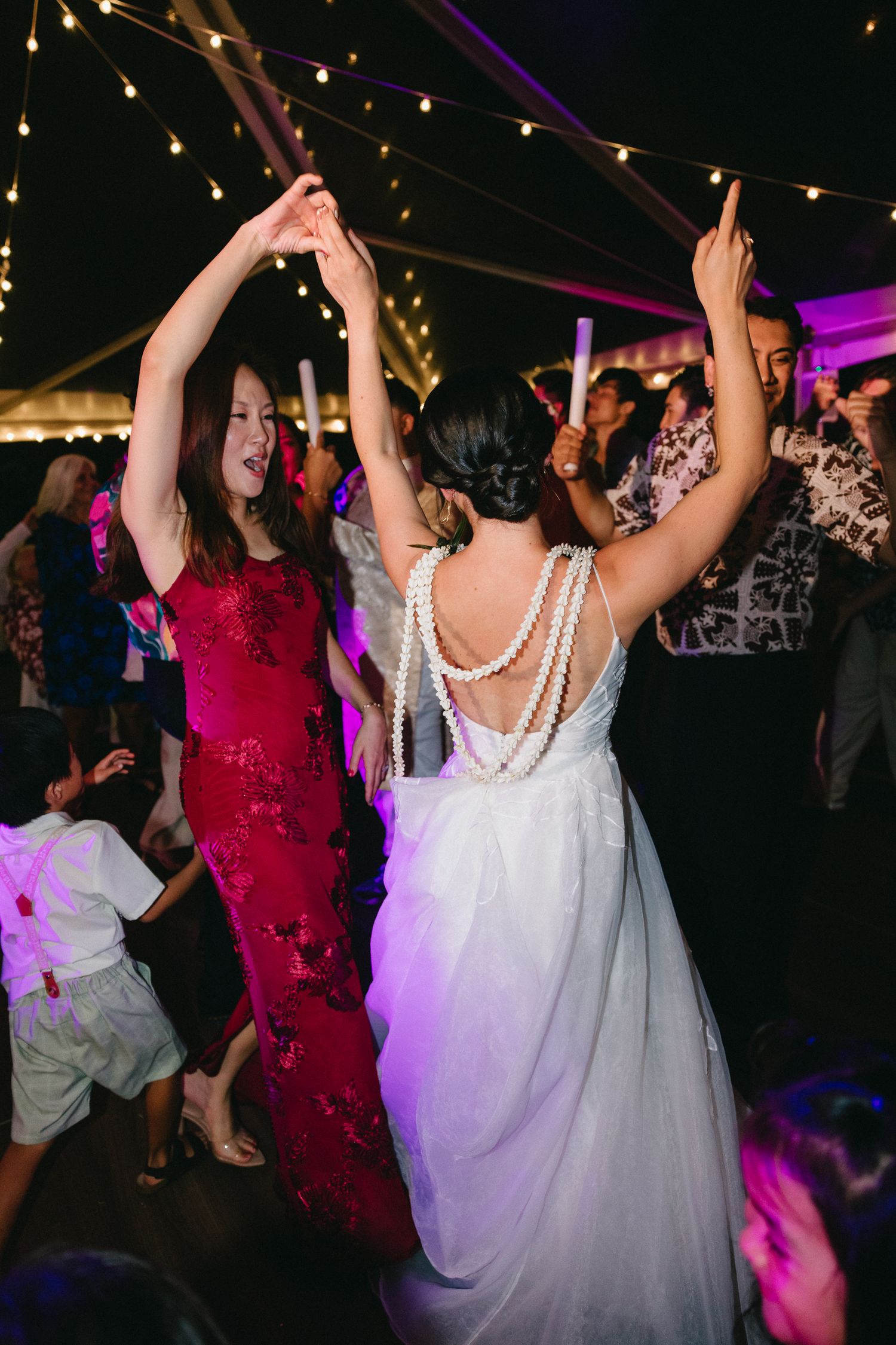 Bride in white gown with pearl back detail dances with guest in red dress at elegant outdoor wedding reception.