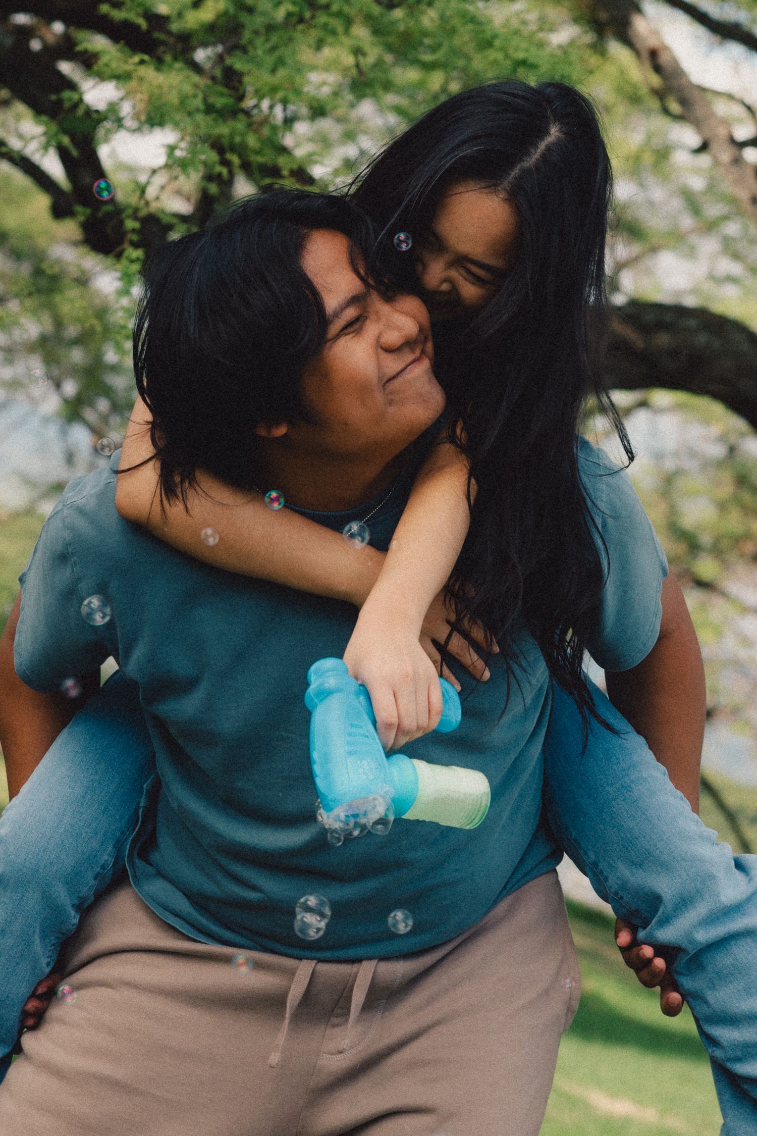 A woman gives a smiling girl a piggyback ride outdoors, both wearing teal tops, with trees in the background.