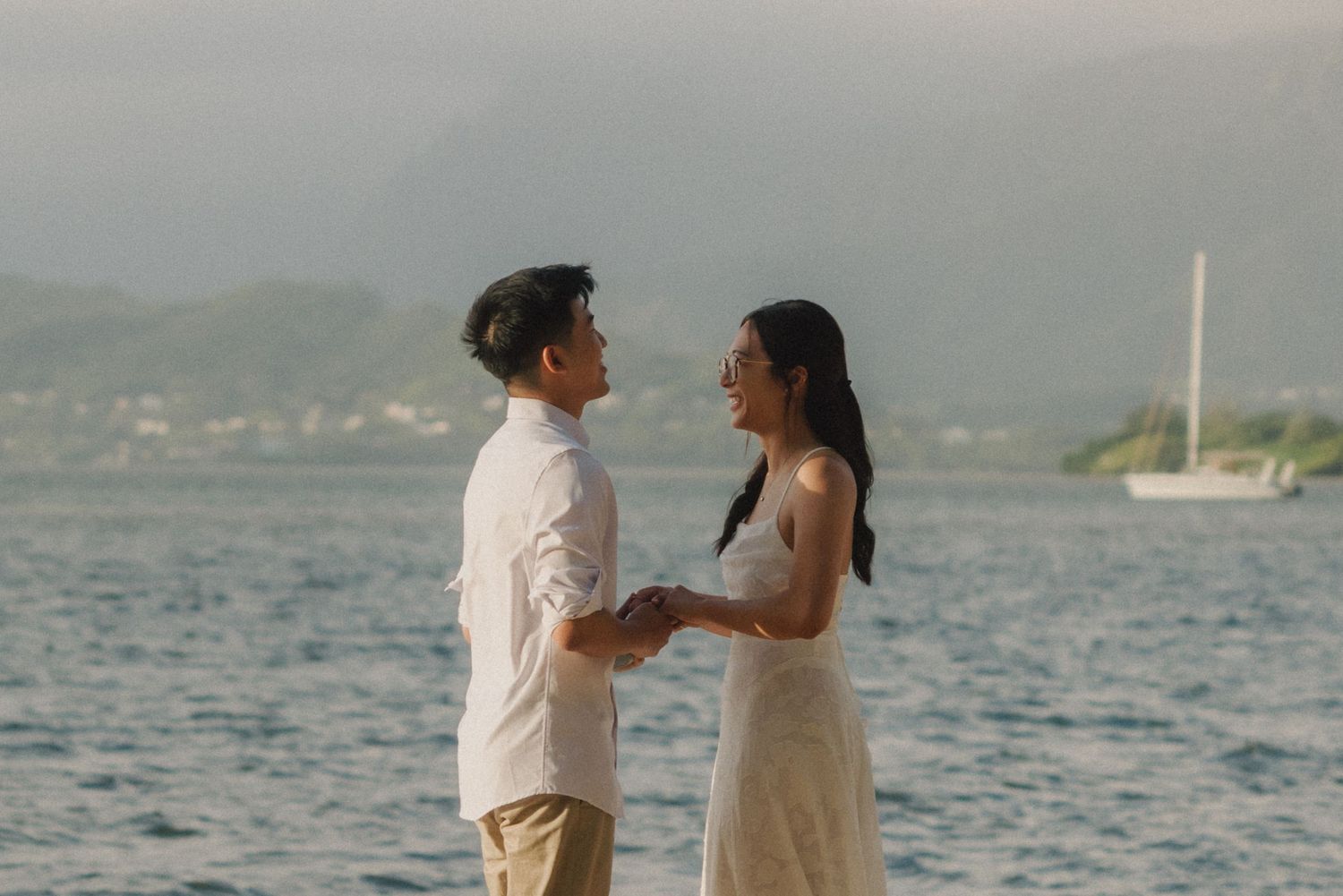 Couple holding hands facing each other by the ocean with mountains and a sailboat in the background at dusk.