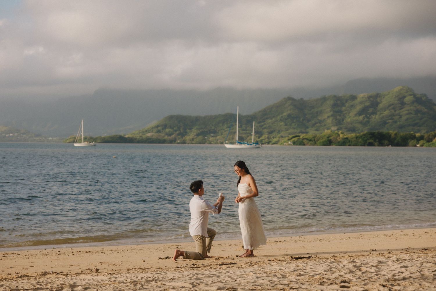Man proposing to woman on a sandy beach in Hawaii with mountains and sailboats in the background.