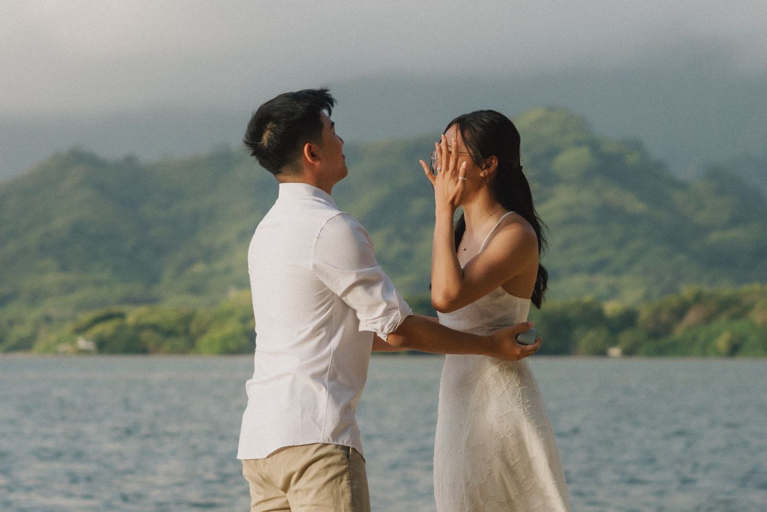 Couple in white outfits sharing a romantic moment by a serene lake with lush green mountains in the background.
