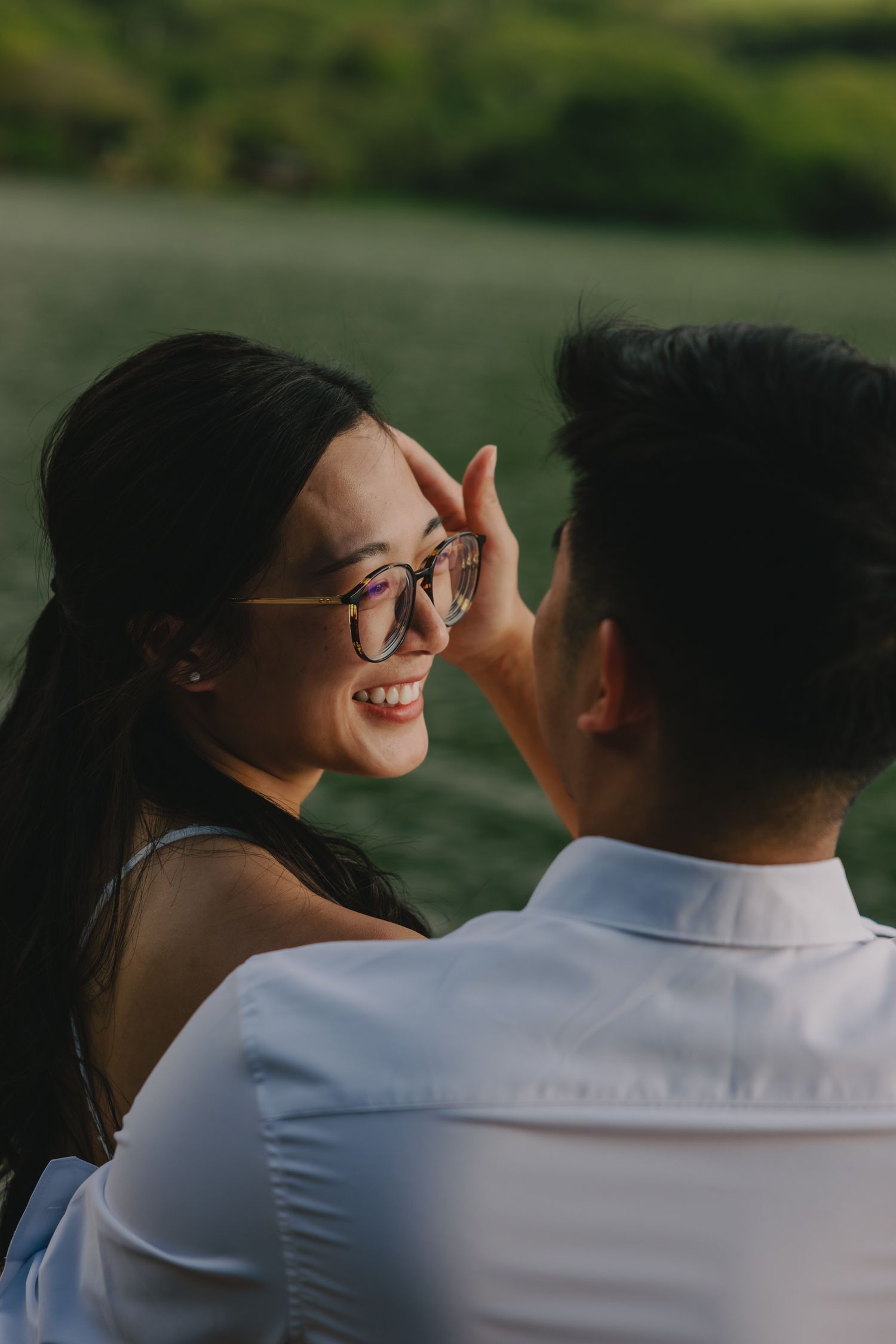 A smiling woman in glasses shares a tender moment with her partner by a serene green lake.